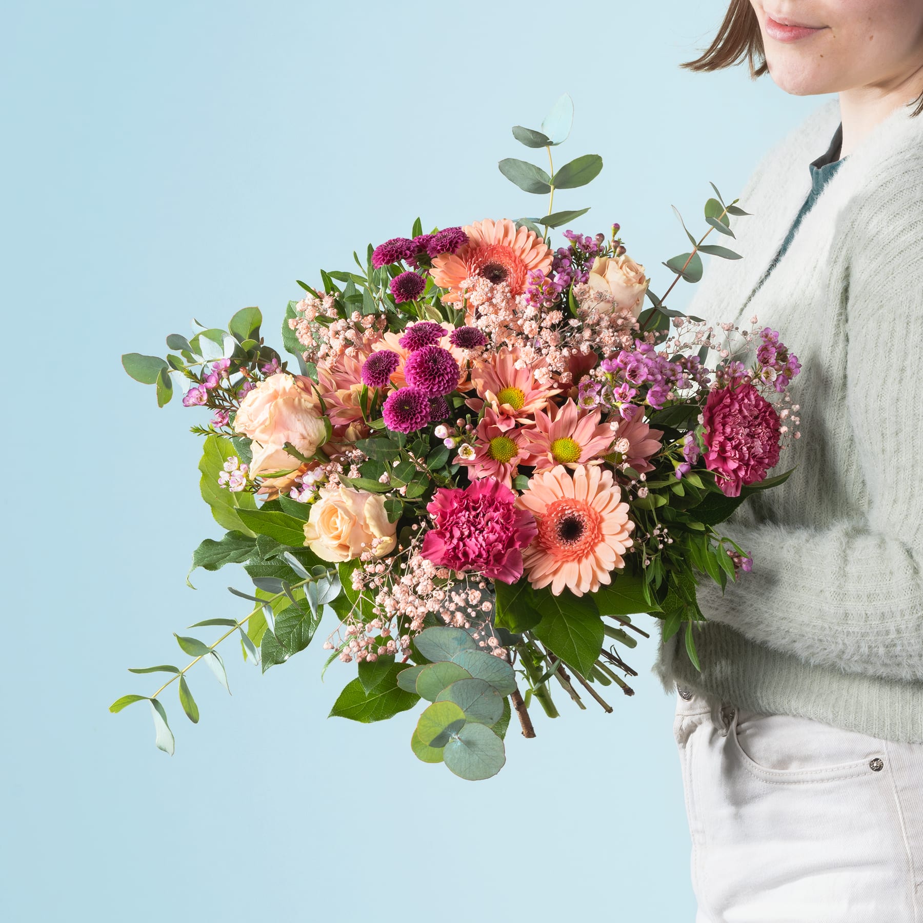 Ein leuchtender Strauß bunter Blumen, darunter Gerbera, Nelken und Eukalyptusblätter, wird von einer Person gehalten, die einen hellen Pullover vor einem schlichten blauen Hintergrund trägt.