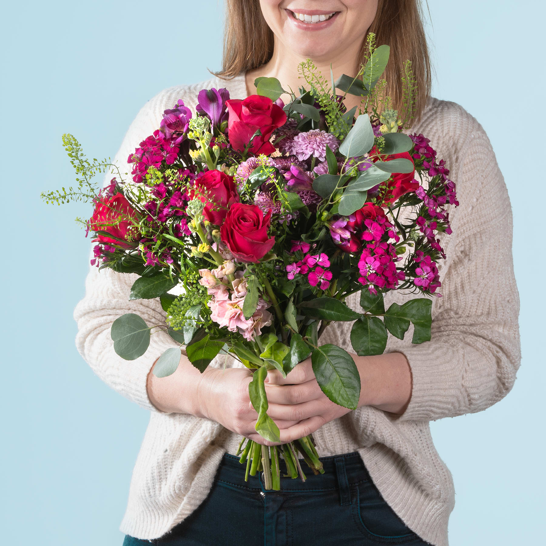 Eine lächelnde Frau hält einen farbenfrohen Strauß roter Rosen, rosa Nelken und anderer bunter Blumen vor einem hellblauen Hintergrund.