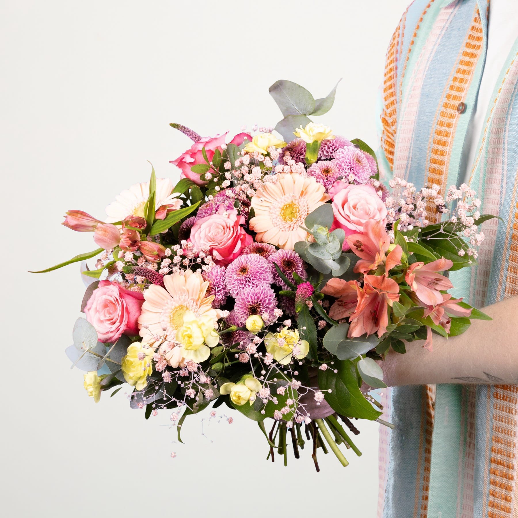 Ein farbenfroher Strauß verschiedener rosa, gelber und weißer Blumen, darunter Rosen, Gänseblümchen und andere Blüten, wird im Vordergrund vor einem schlichten weißen Hintergrund gehalten.