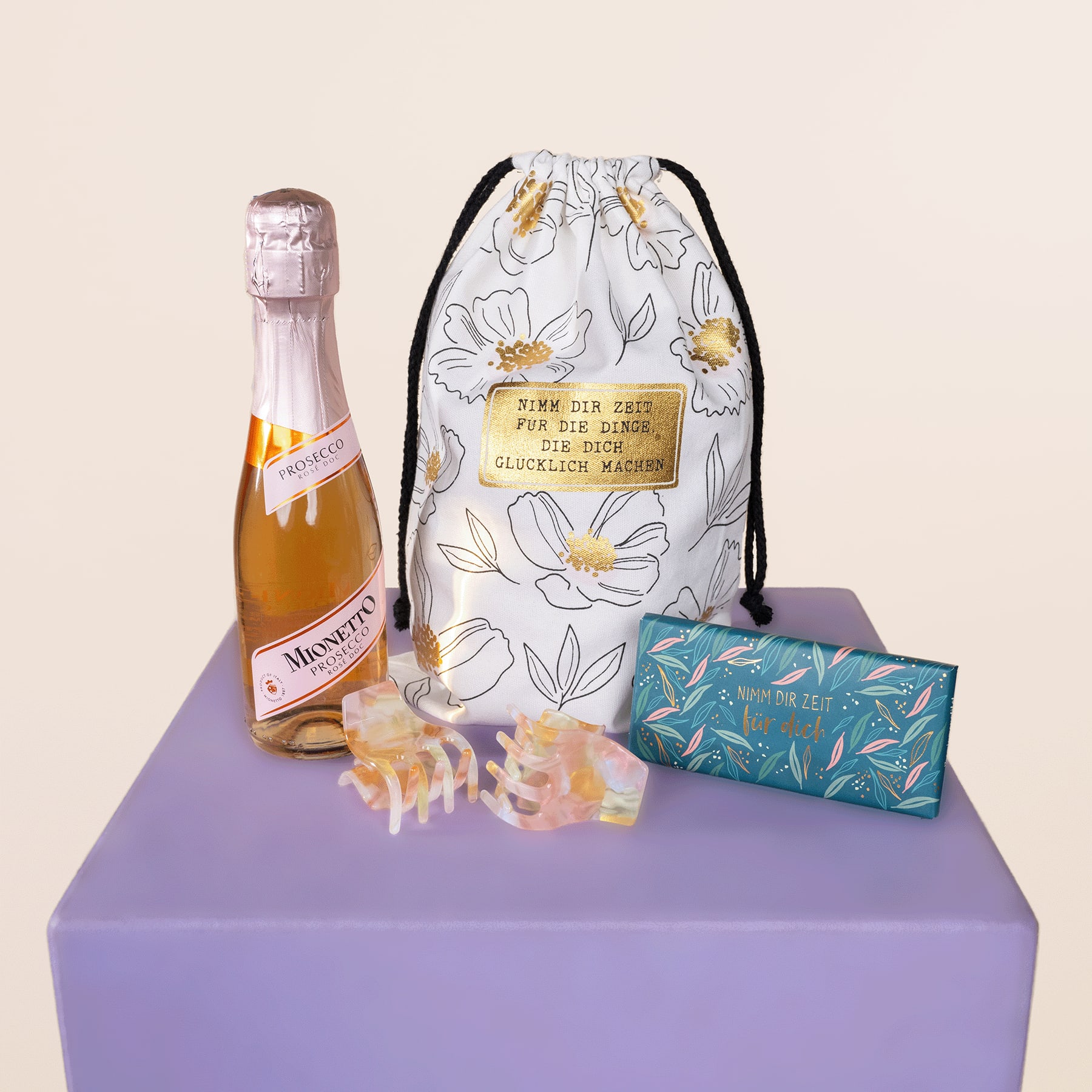 Eine Flasche Sekt, ein Beutel mit Blumenmuster und eine Schachtel Pralinen sind auf einem lavendelfarbenen Podest vor schlichtem Hintergrund arrangiert.