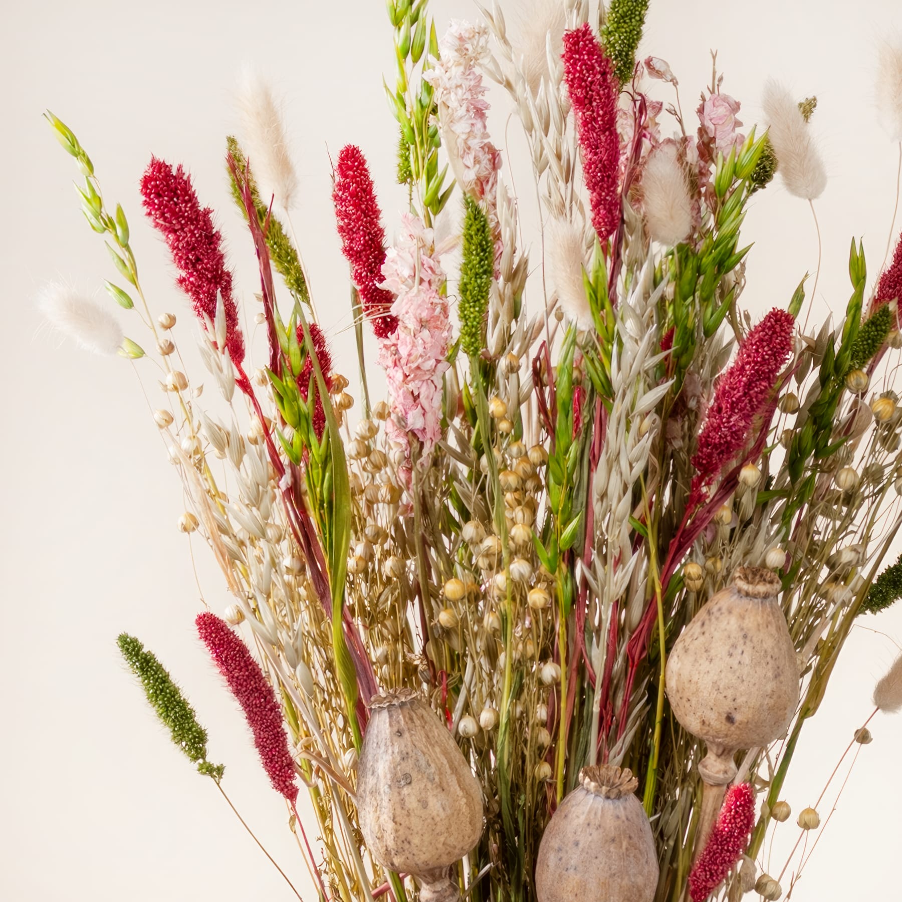 Ein farbenfroher Strauß verschiedener getrockneter Blumen und Gräser, darunter rote und rosa Blüten, grünes Laub und strukturierte Samenkapseln, arrangiert in einer rustikalen Vase vor einem schlichten Hintergrund.