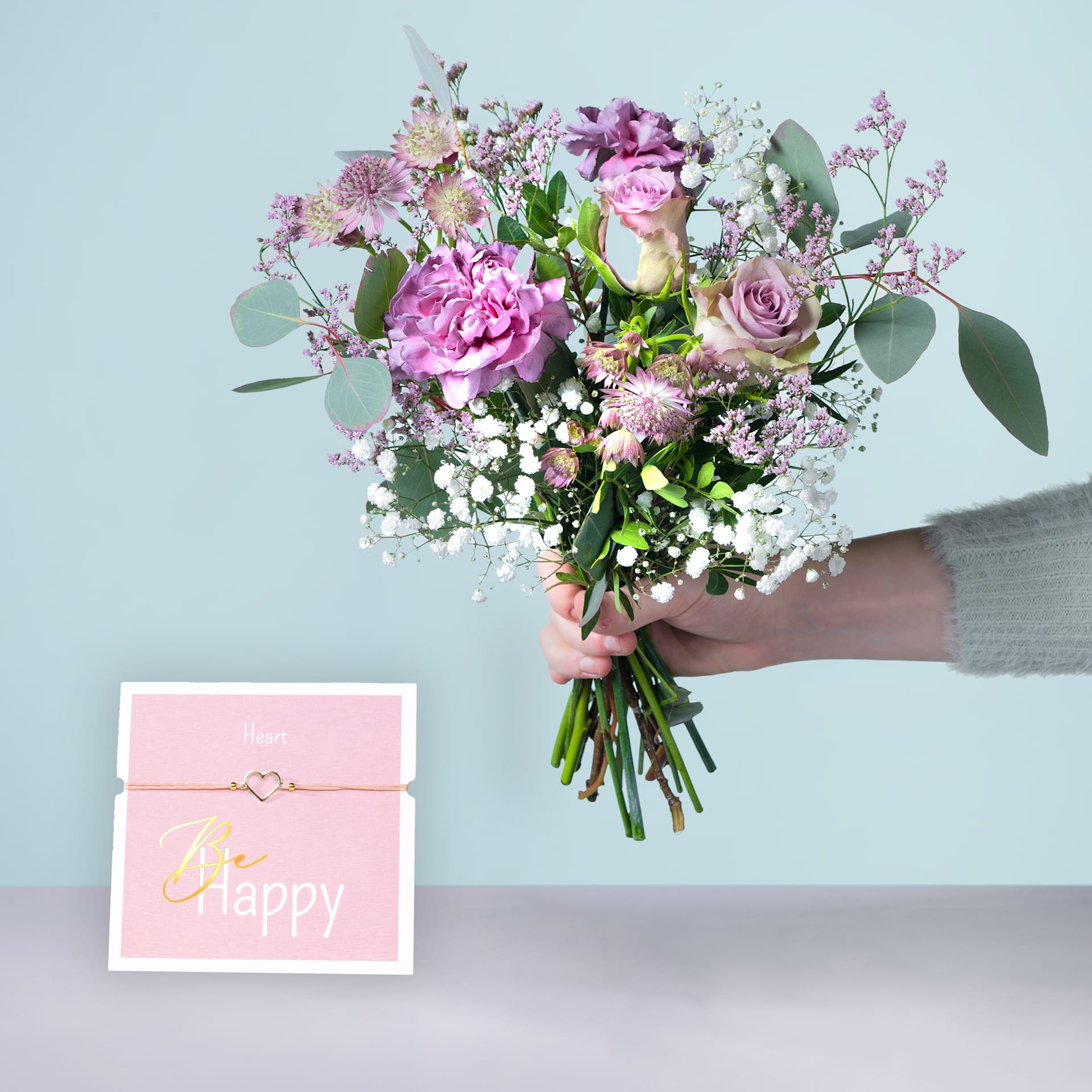 Eine Person hält einen farbenfrohen Strauß rosa und weißer Blumen, darunter Pfingstrosen und Rosen, in den Händen, begleitet von einer rosa Geschenkbox mit der Aufschrift „Happy“.
