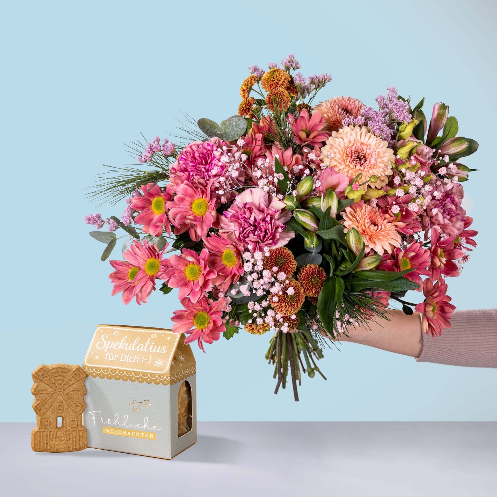 Ein farbenfroher Strauß rosa, orangefarbener und violetter Blumen, darunter Dahlien und Chrysanthemen, wird vor einer hölzernen Geschenkbox vor einem hellblauen Hintergrund gehalten.