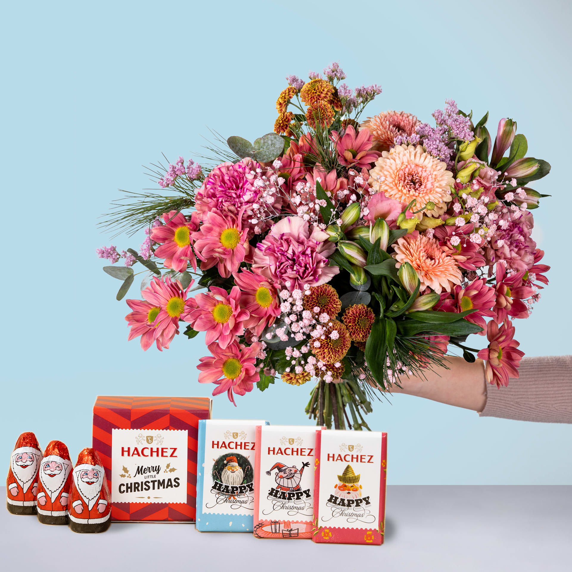 Im Vordergrund steht ein farbenfroher Strauß rosa, orangefarbener und violetter Blumen, im Hintergrund mehrere kleine Weihnachtsmannfiguren und Geschenkboxen mit der Aufschrift „Hachez“.