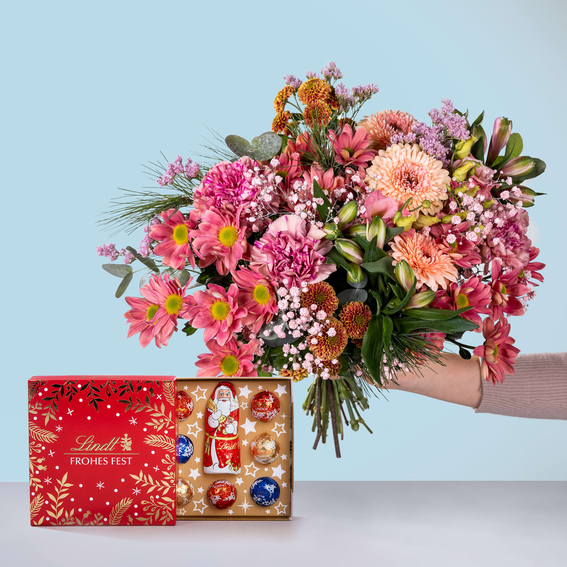 Ein farbenfroher Strauß rosa, orangefarbener und violetter Blumen, darunter Dahlien und Chrysanthemen, wird vor einer roten Geschenkbox mit festlichem Feiertagsdesign präsentiert.