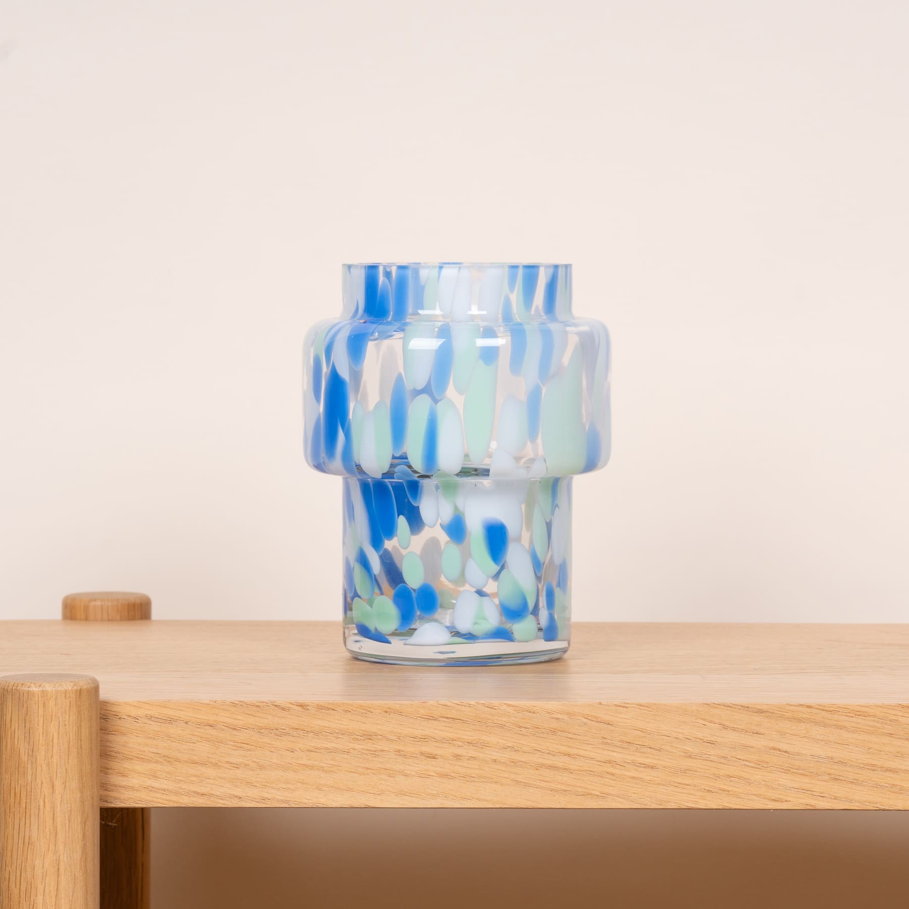 Eine bunte Glasvase mit einem wirbelnden Muster aus blauen und weißen Farbtönen steht auf einem Holztisch vor einem schlichten beigen Hintergrund.
