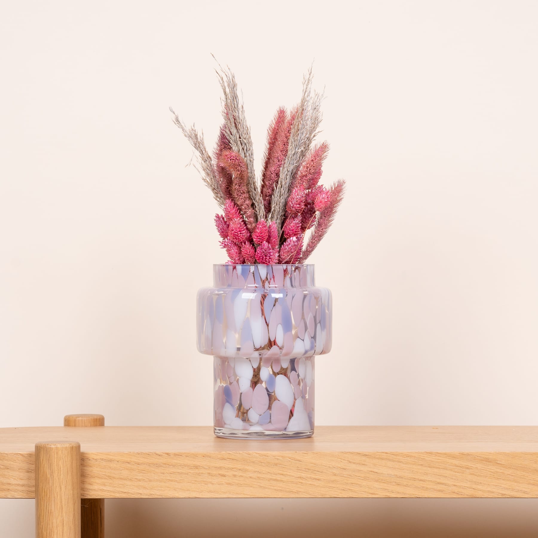 Eine Glasvase mit leuchtend rosa und weißen Trockenblumen steht auf einem Holztisch vor einem schlichten beigen Hintergrund.