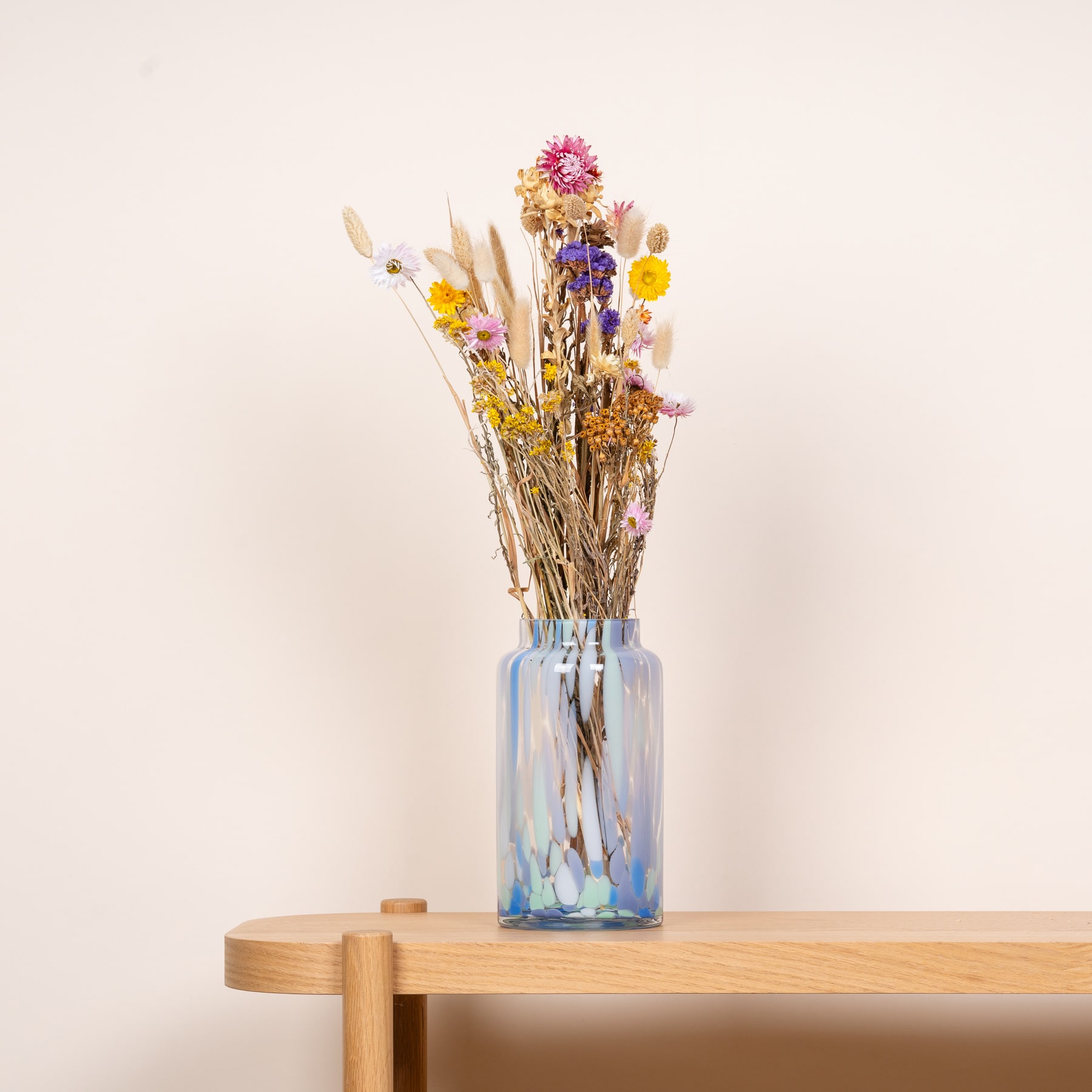 Ein Strauß leuchtender Trockenblumen in einer Glasvase steht auf einem Holztisch vor einem schlichten, hellen Hintergrund.