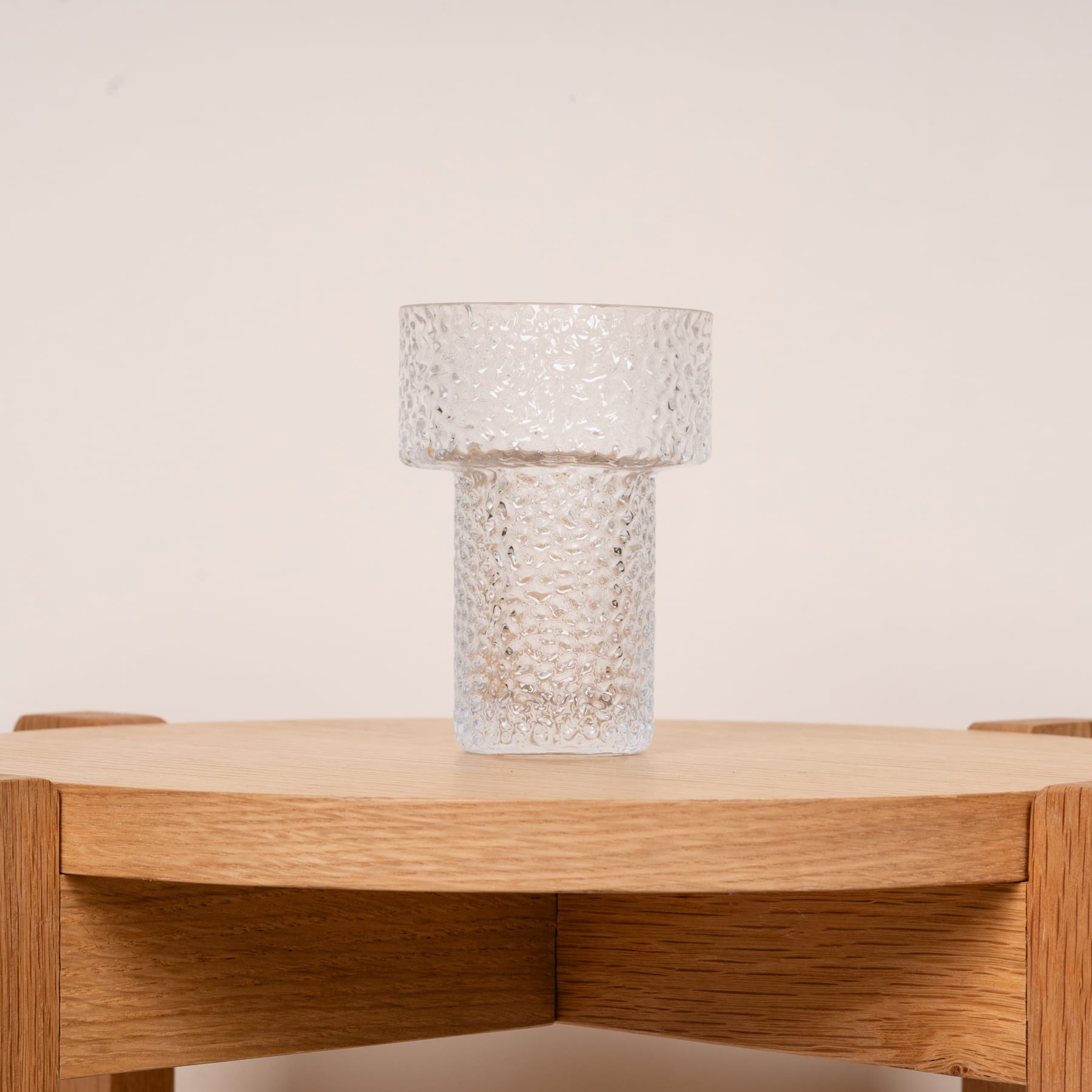 Eine Glaslampe mit strukturiertem, gemustertem Schirm steht auf einem Holztisch oder Regal in einer minimalistischen Umgebung in neutralen Tönen.