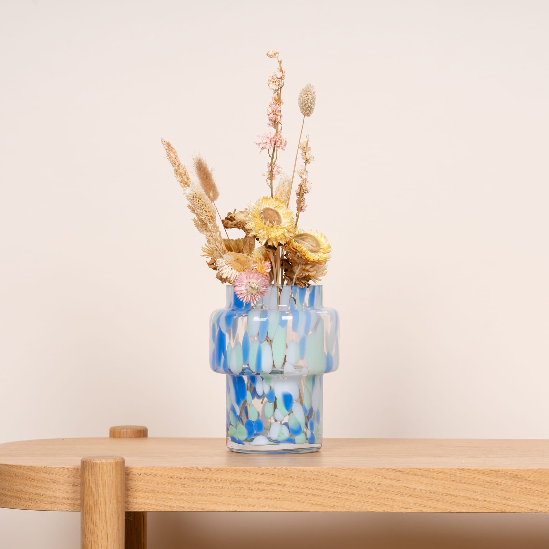 Ein farbenfroher Strauß getrockneter Blumen und Gräser wird in einer dekorativen Glasvase präsentiert, die auf einer Holzoberfläche vor einem schlichten Hintergrund ruht.