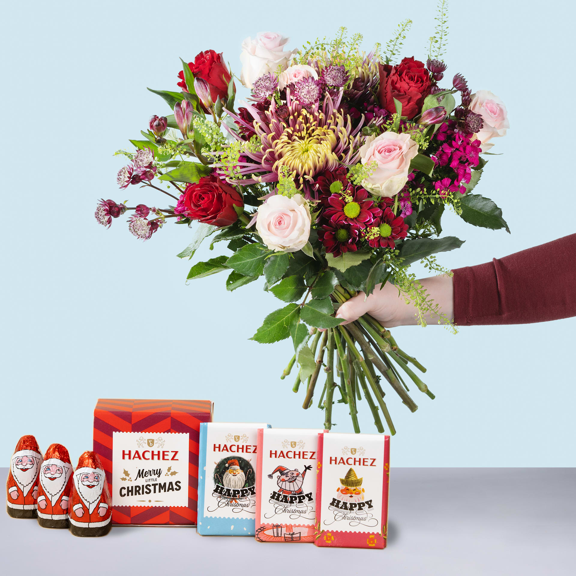 Eine Person hält einen farbenfrohen Strauß roter Rosen, rosa Pfingstrosen und gelber Chrysanthemen in den Händen, begleitet von mehreren festlichen Weihnachtsmannfiguren aus Schokolade und Geschenkboxen mit Feiertagsmotiven.