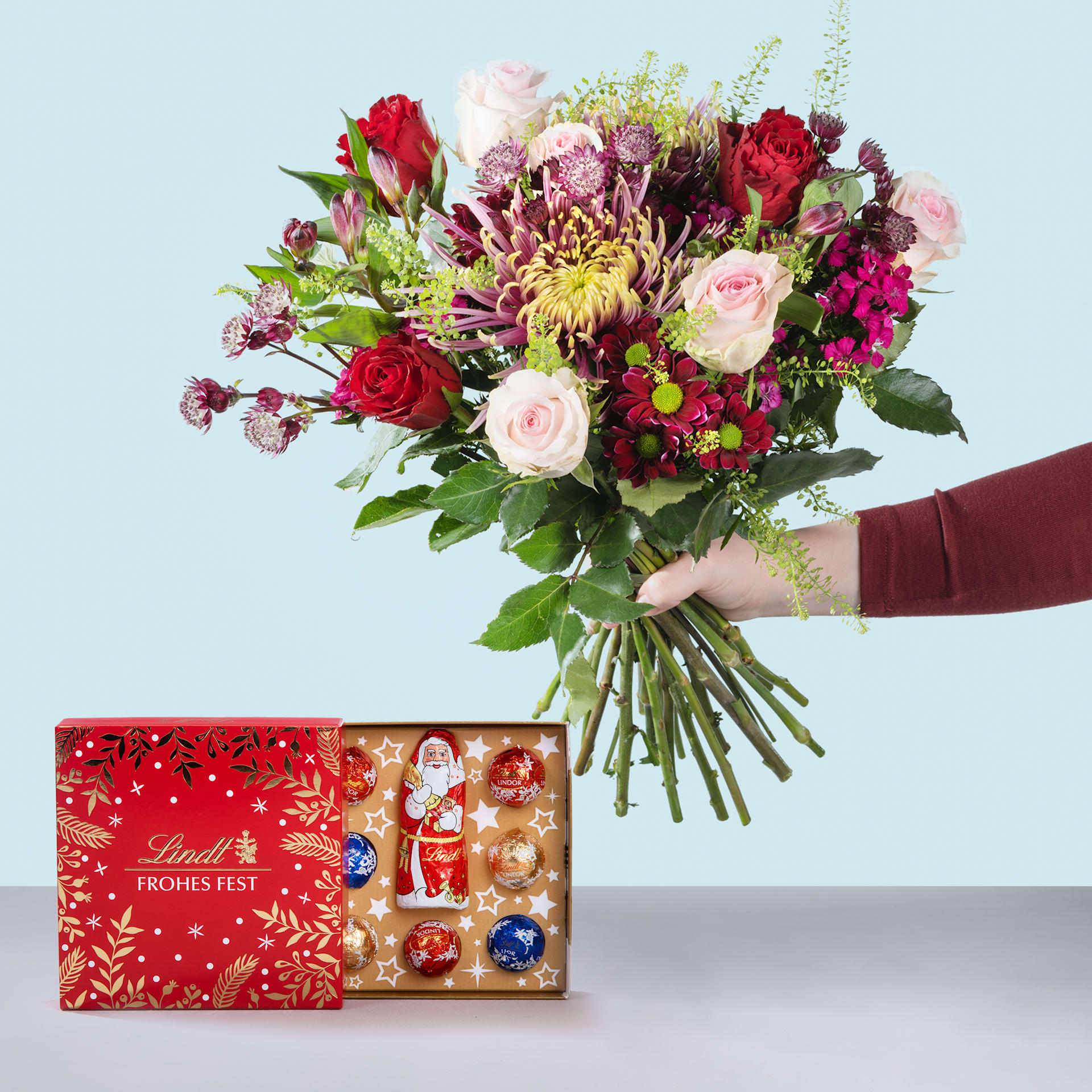 Ein leuchtender Strauß roter Rosen, rosa Pfingstrosen und anderer bunter Blumen steht vor einer roten Geschenkbox mit festlichem Feiertagsdesign.