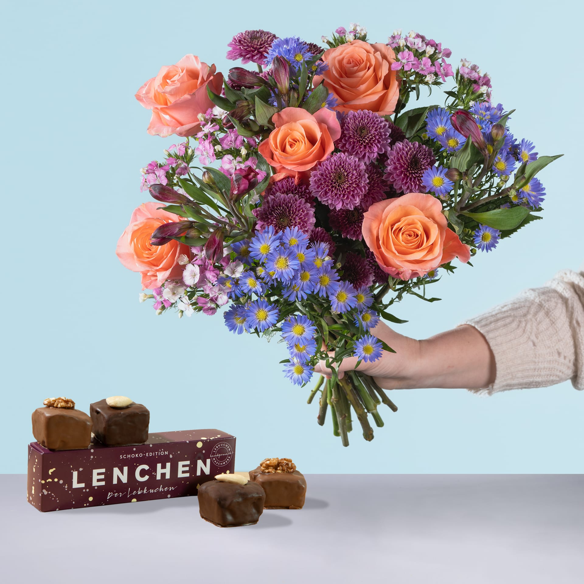 Eine Person hält einen leuchtenden Strauß bunter Blumen, darunter Rosen, Chrysanthemen und Gänseblümchen, in den Händen. Im Vordergrund ist vor einem hellblauen Hintergrund eine Schachtel Pralinen mit der Aufschrift „Lenchen“ zu sehen.