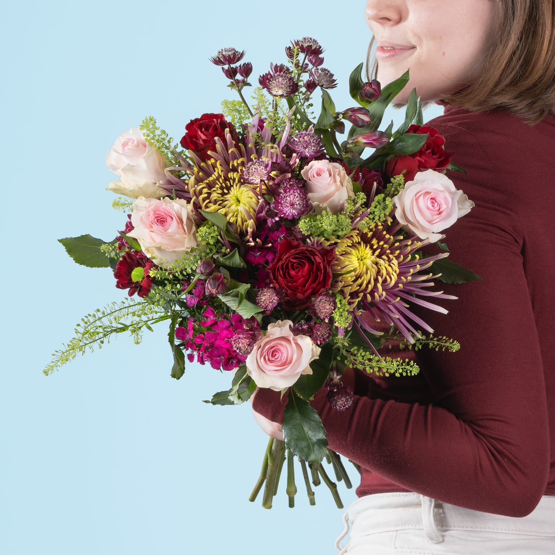 Ein leuchtender Strauß bunter Blumen, darunter Rosen, Dahlien und Chrysanthemen, wird von einer Person gehalten, die einen burgunderfarbenen Pullover vor einem hellblauen Hintergrund trägt.