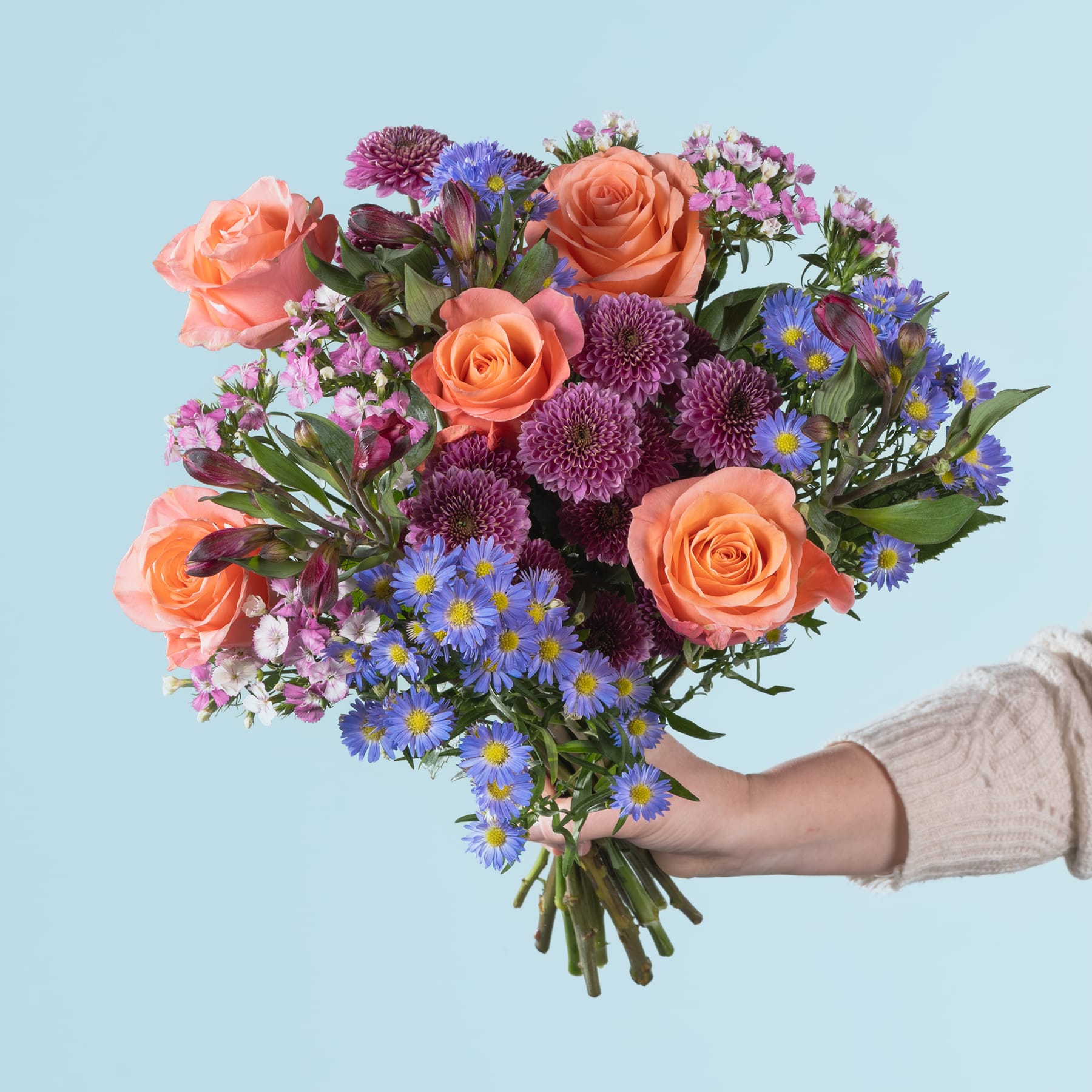Ein leuchtender Strauß bunter Blumen, darunter orangefarbene Rosen, violette Chrysanthemen und blaue Gänseblümchen, wird vor einem hellblauen Hintergrund in den Händen einer Person gehalten.
