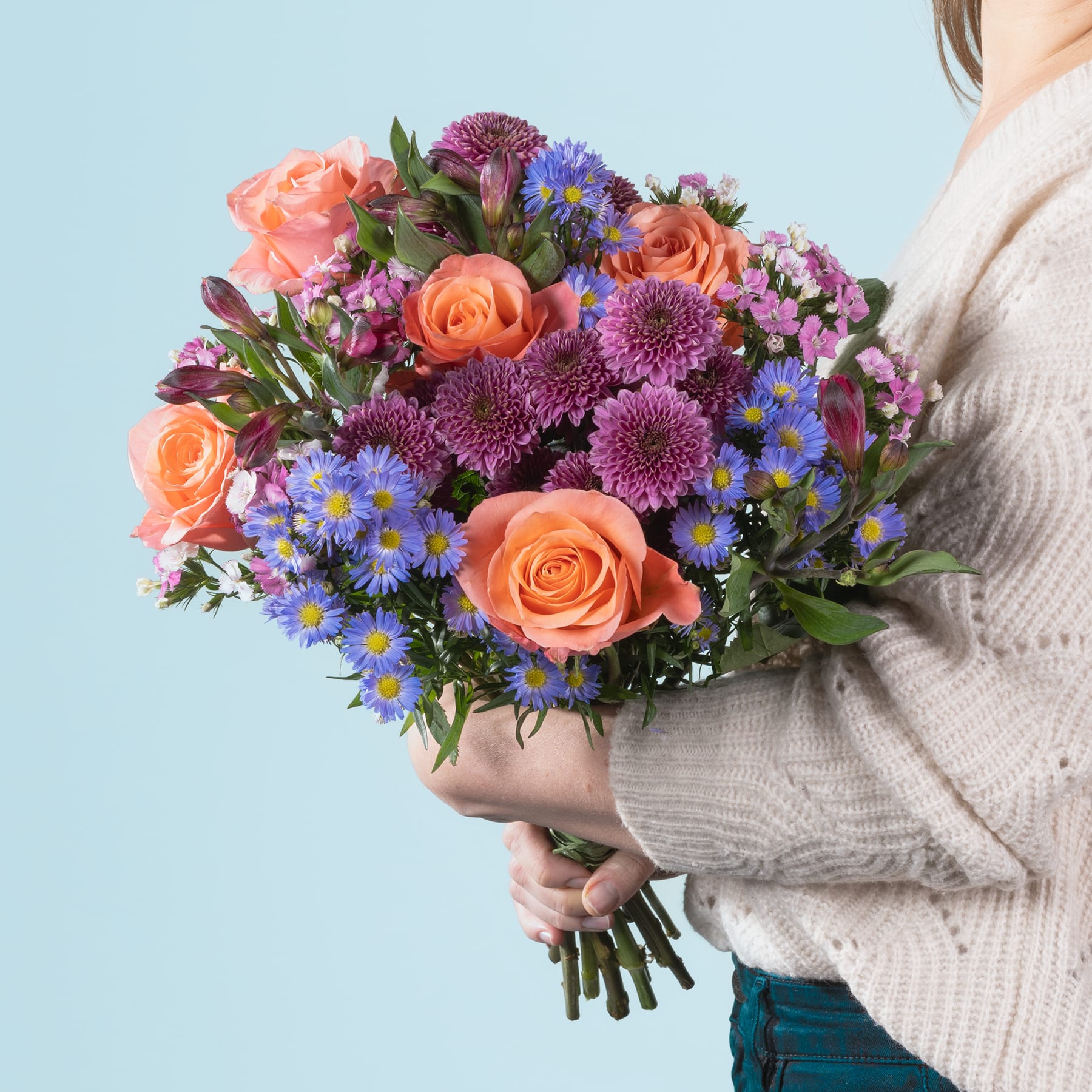 Ein leuchtender Strauß bunter Blumen, darunter orangefarbene Rosen, violette Chrysanthemen und blaue Gänseblümchen, wird in den Händen einer Person gehalten, die einen grauen Pullover vor einem hellblauen Hintergrund trägt.