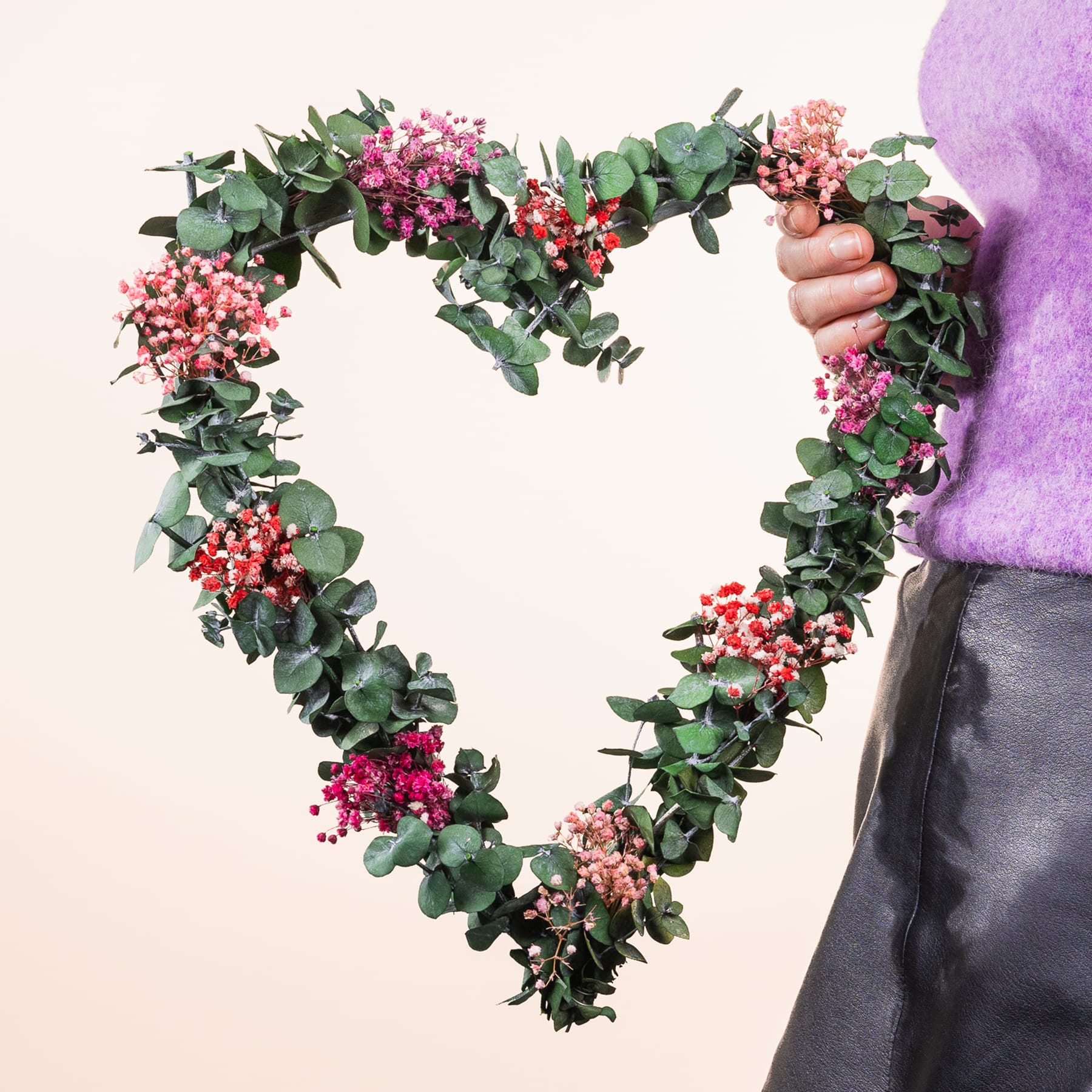 Ein herzförmiger Kranz aus üppigem Grün und leuchtend rosa Blumen wird vor einer Person gehalten, die einen lila Pullover vor einem schlichten Hintergrund trägt.