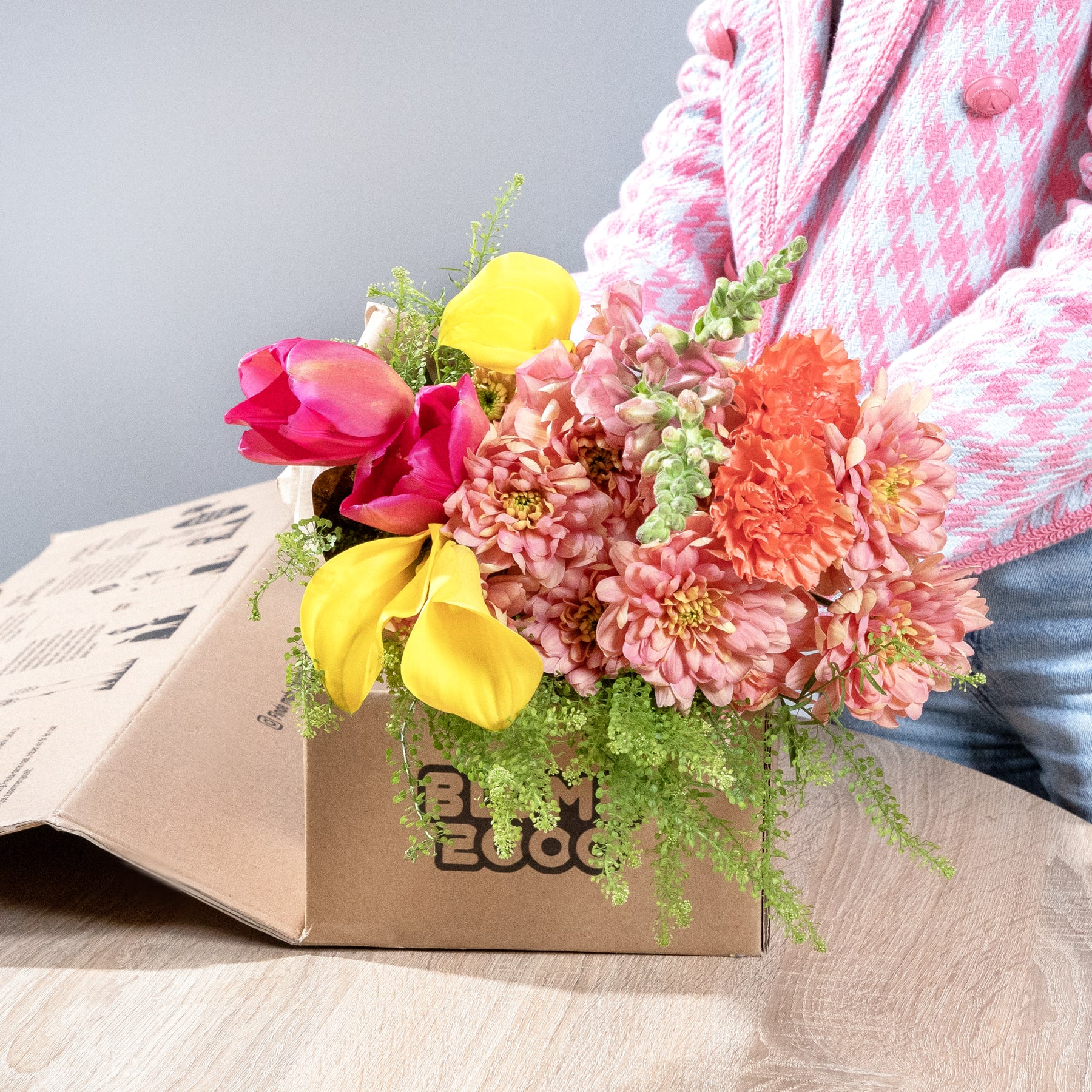 Ein leuchtender Strauß bunter Blumen, darunter rote Tulpen, gelbe Narzissen und rosa Chrysanthemen, steht in einem Karton auf einer Holzoberfläche, im Hintergrund ist ein rosa-weiß karierter Stoff zu sehen.