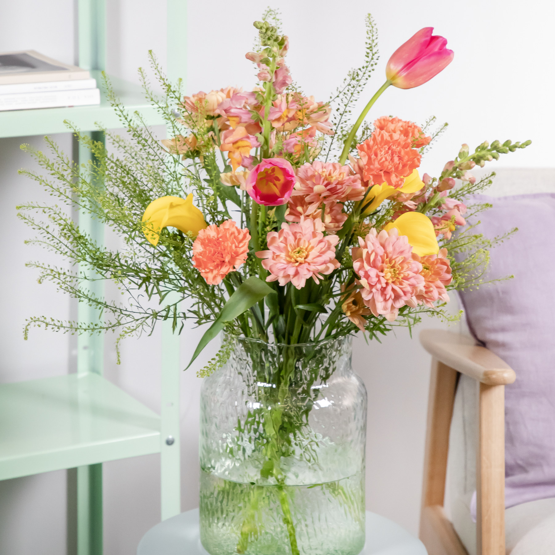 Ein leuchtender Strauß bunter Blumen, darunter Tulpen, Gänseblümchen und Nelken, wird in einer Glasvase vor dem Hintergrund eines Holzstuhls und eines grünen Regals präsentiert.