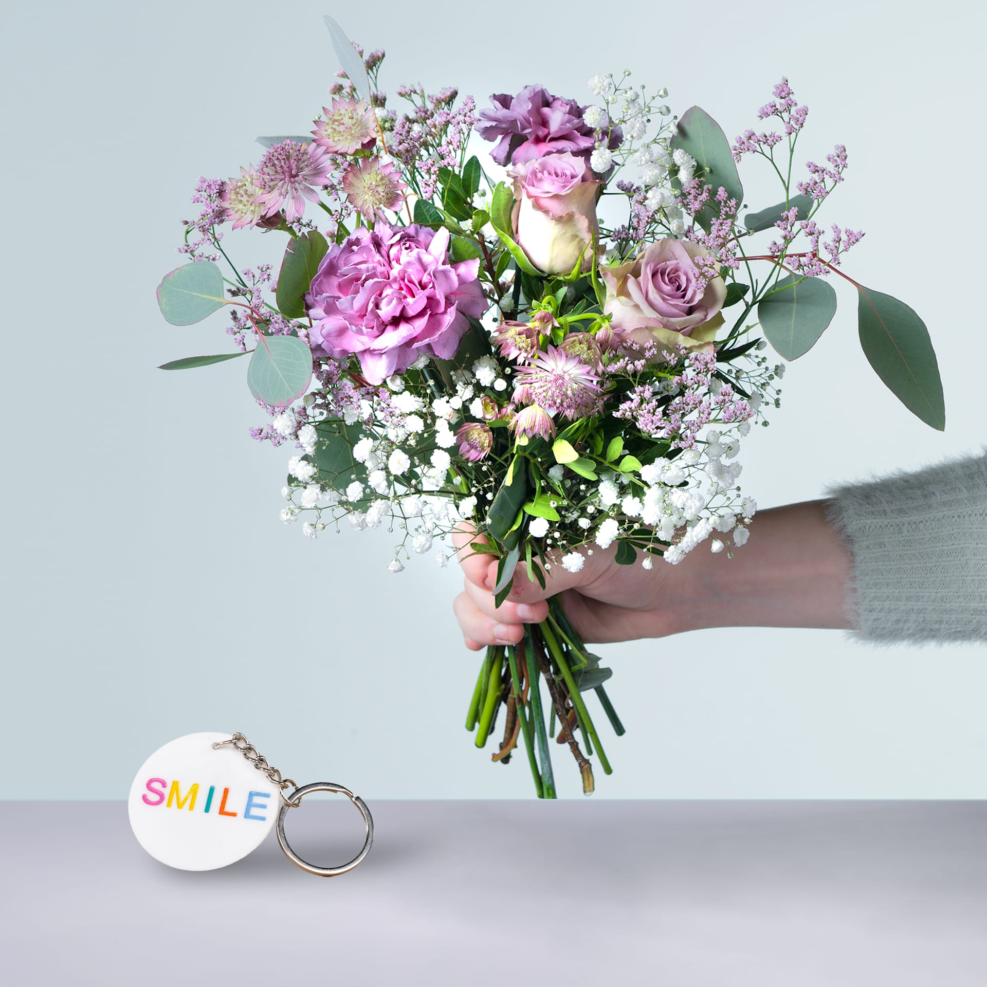 Eine Person hält einen farbenfrohen Strauß rosa und weißer Blumen, darunter Rosen und Pfingstrosen, in den Händen, begleitet von einem Schlüsselanhänger mit dem Wort „SMILE“ auf grauem Hintergrund.