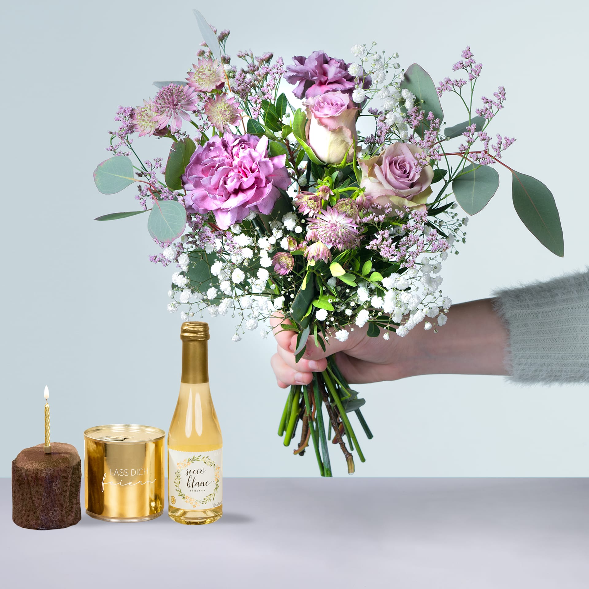 Eine Person hält einen Strauß leuchtend rosa und weißer Blumen, darunter Rosen und Pfingstrosen, in den Händen. Im Hintergrund stehen auf einem Tisch eine Flasche Champagner und eine Kerze.