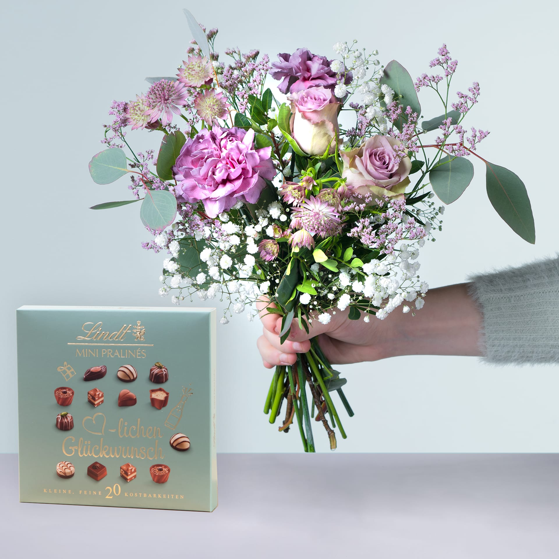 Eine Person hält einen Strauß leuchtend rosa und weißer Blumen, darunter Rosen und Pfingstrosen, in den Händen, begleitet von einer Schachtel verschiedener Pralinen auf einem hellen Hintergrund.