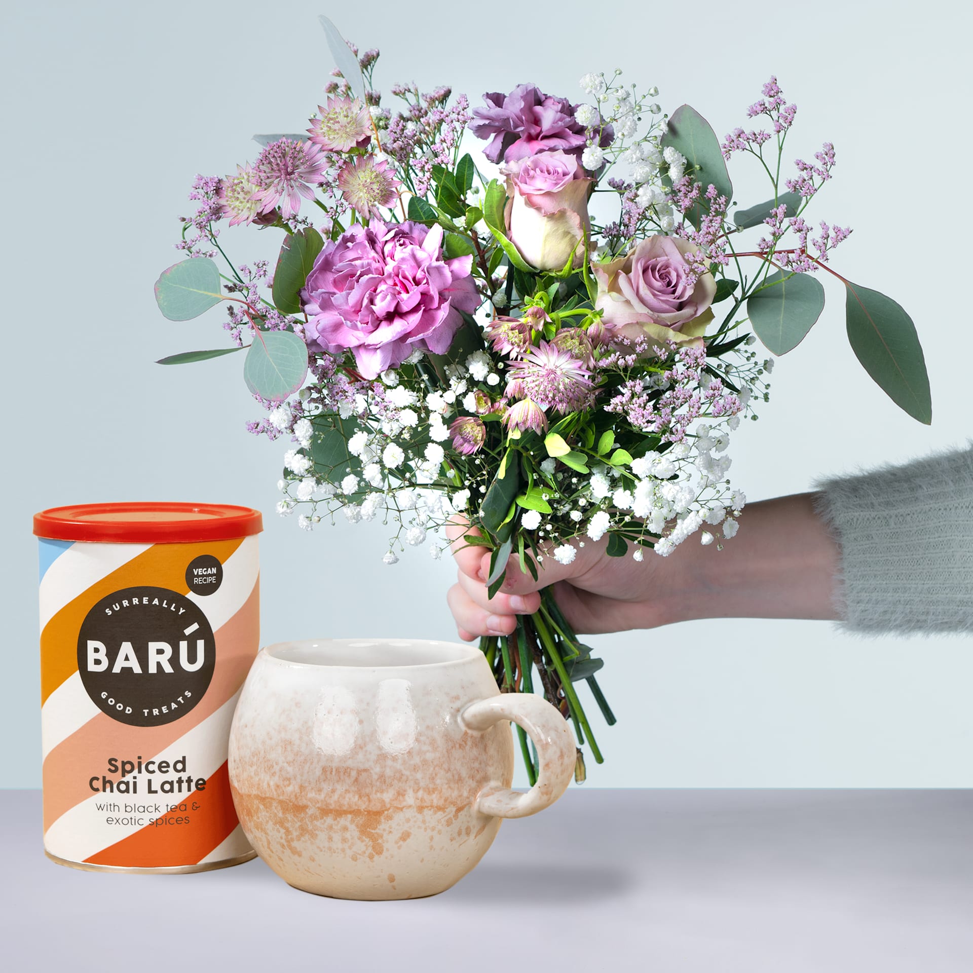 Ein Strauß leuchtend rosa und weißer Blumen, darunter Rosen und Pfingstrosen, wird in einer Keramiktasse neben einer Dose Chai Latte mit Baru-Gewürzen auf einem hellen Hintergrund präsentiert.