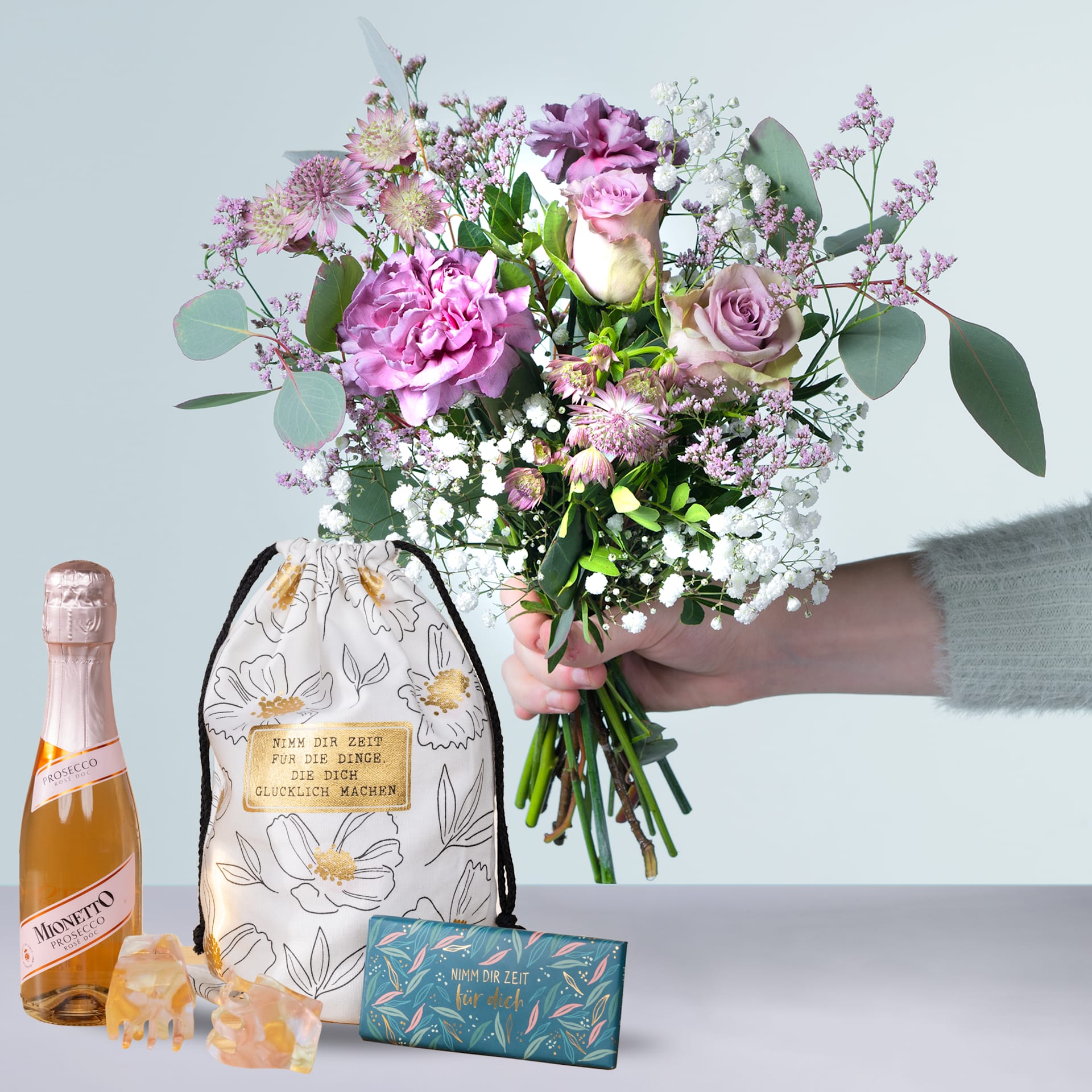 Eine Person hält einen farbenfrohen Strauß rosa und weißer Blumen, darunter Rosen und Pfingstrosen, in den Händen, begleitet von einer Flasche Champagner und einer dekorativen Schachtel auf einem hellen Hintergrund.