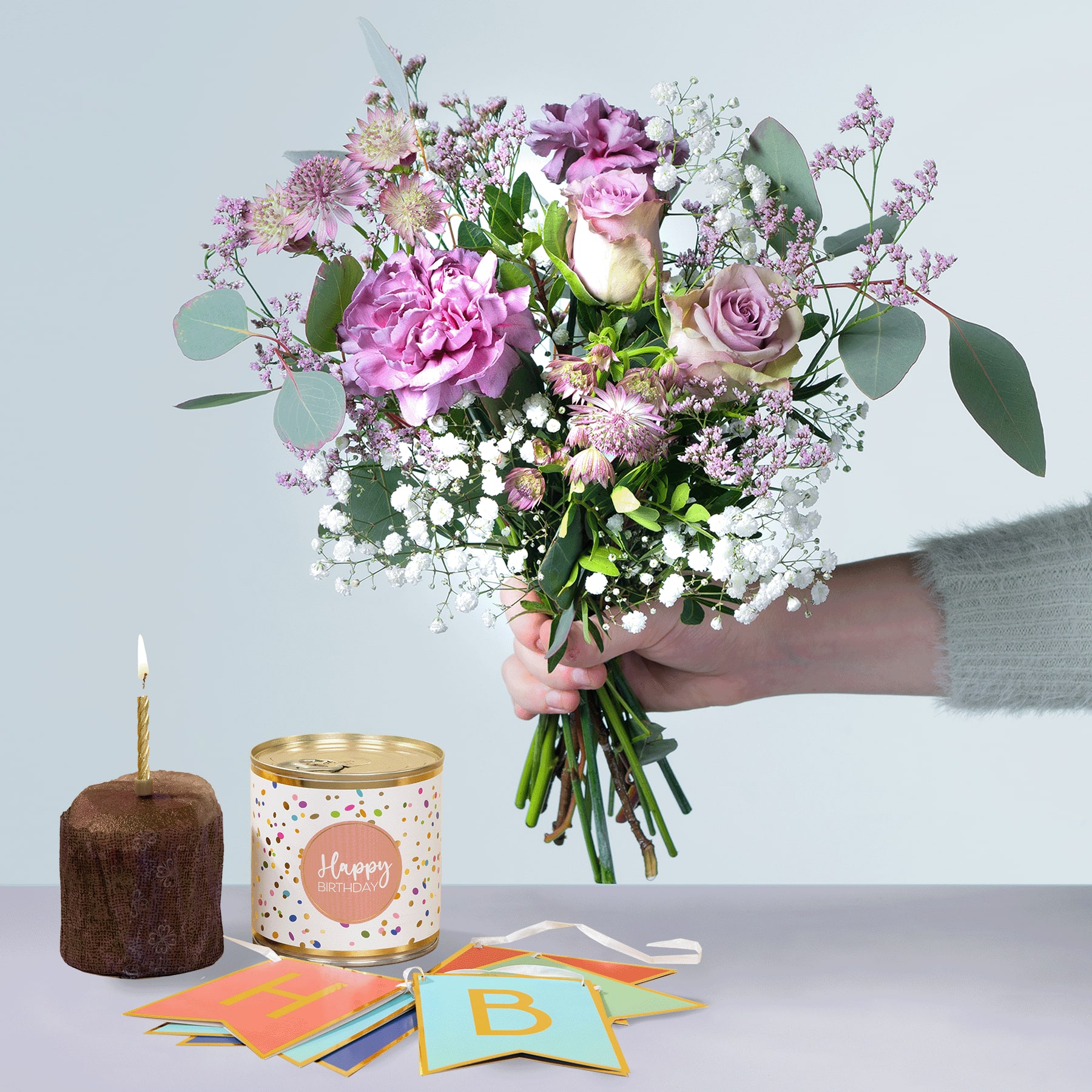 Eine Person hält einen farbenfrohen Strauß rosa und weißer Blumen, darunter Rosen und Pfingstrosen, in den Händen, begleitet von einer Kerze, einer Geschenkbox und einer Geburtstagskarte auf einem hellen Hintergrund.