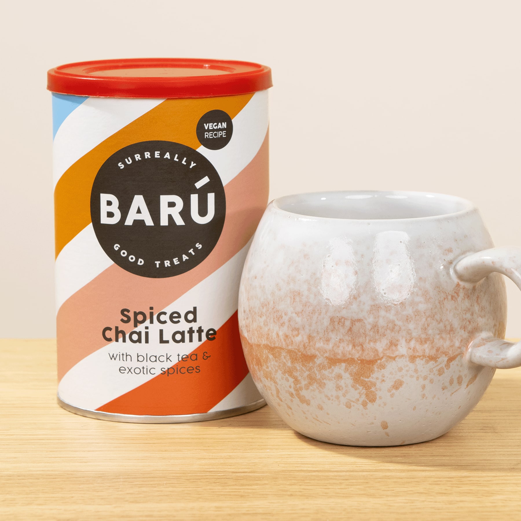 Ein zylindrischer Behälter mit der Aufschrift „BARU Spiced Chai Latte“ steht neben einer weißen Keramiktasse auf einer Holzoberfläche.