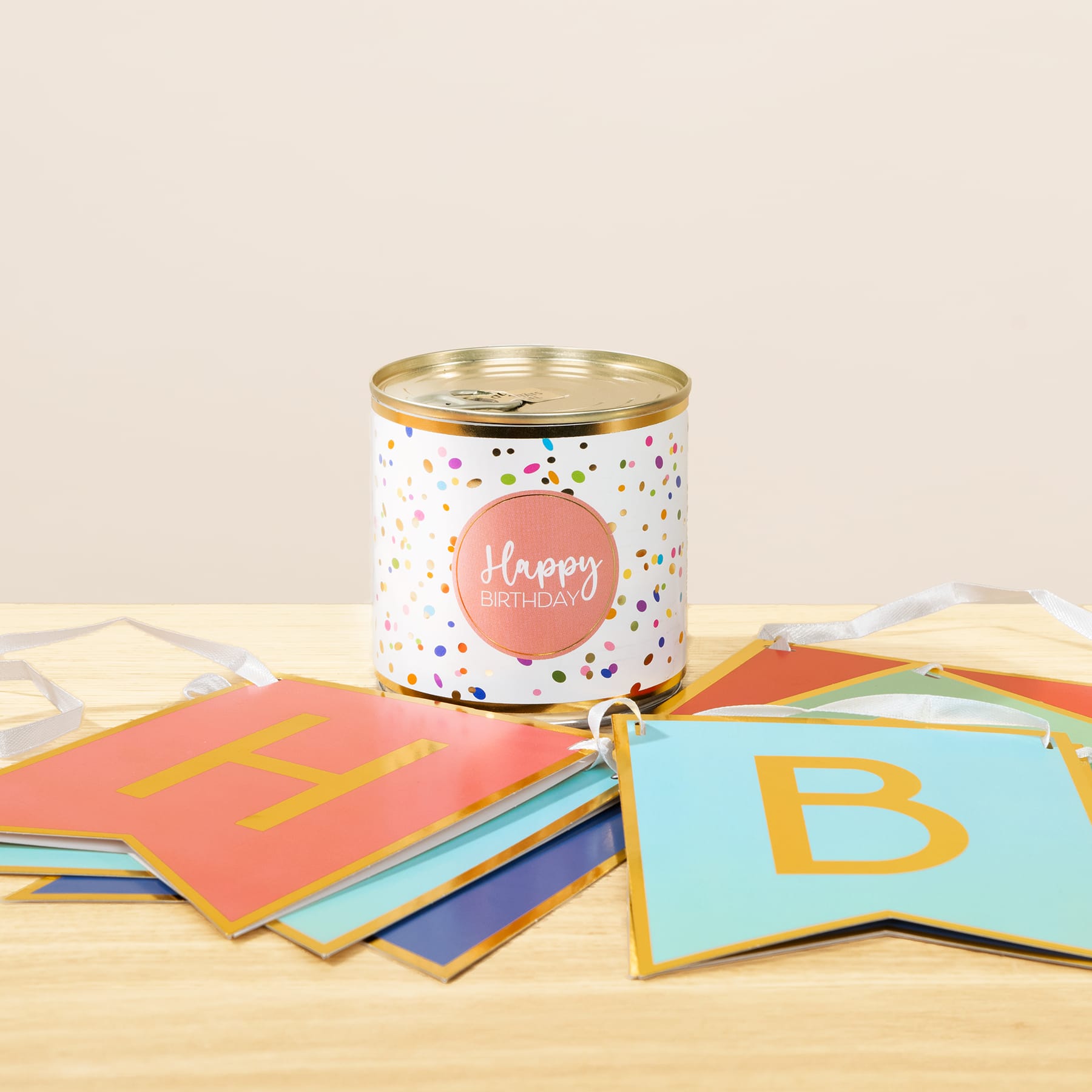Eine bunte Blechdose mit dem Etikett „Happy Birthday“ steht auf einem Stapel farbenfroher Papierkarten auf einer Holzoberfläche.