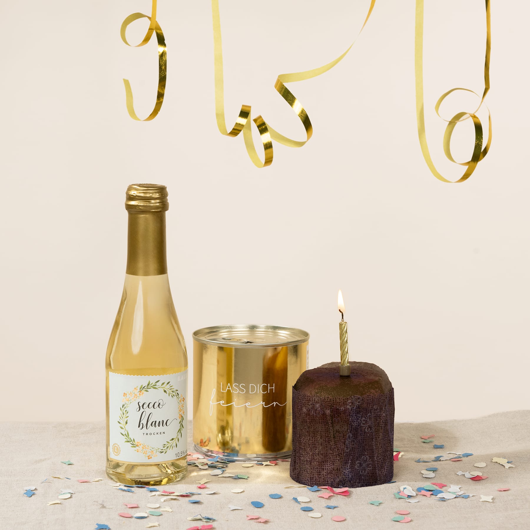 Eine Flasche Weißwein, eine goldene Blechdose und eine brennende Geburtstagskerze auf einem Tisch, umgeben von buntem Konfetti und sich kräuselnden Bändern.