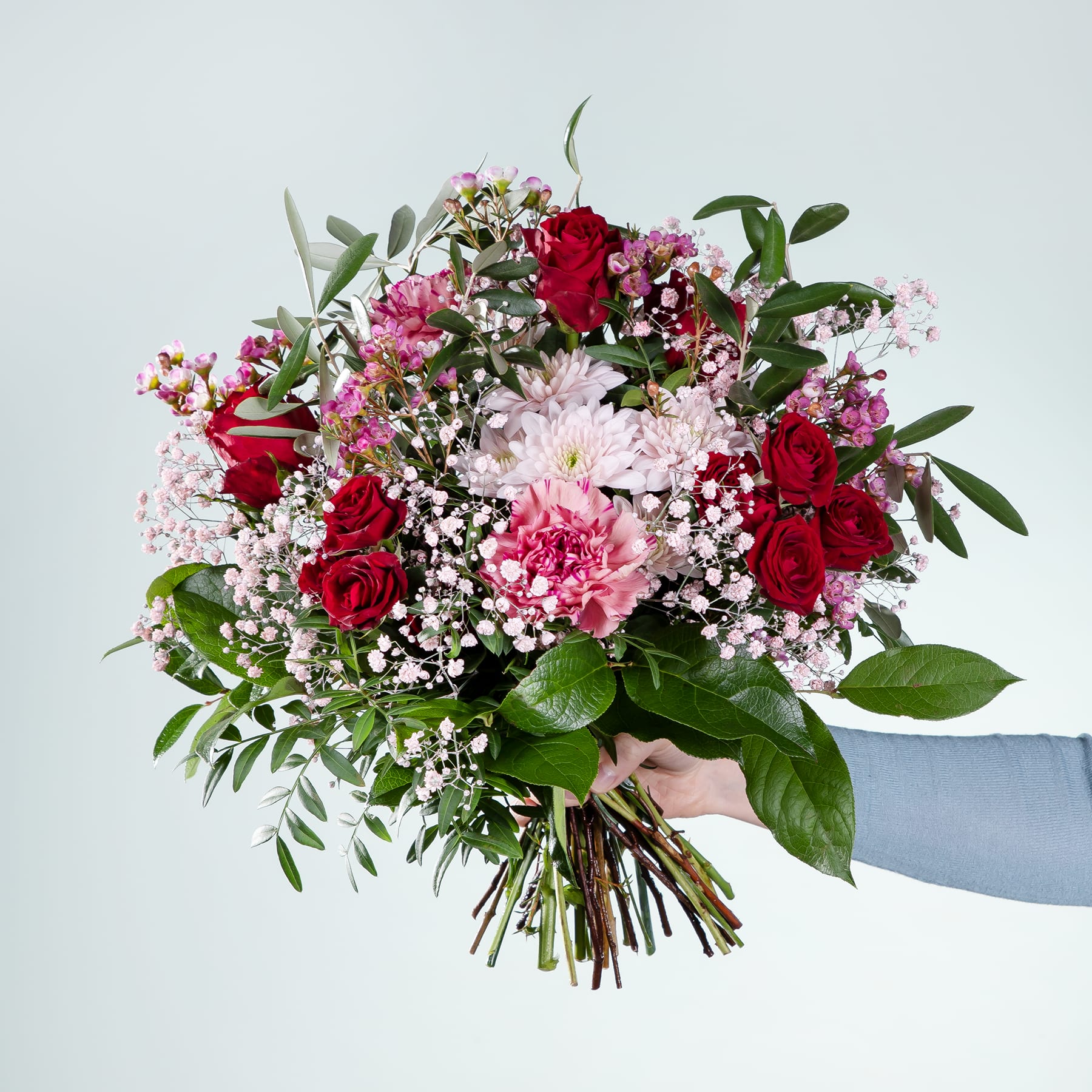 Ein leuchtender Strauß aus roten Rosen, rosa Nelken und weißem Schleierkraut, den eine Person vor einem schlichten Hintergrund in den Händen hält.