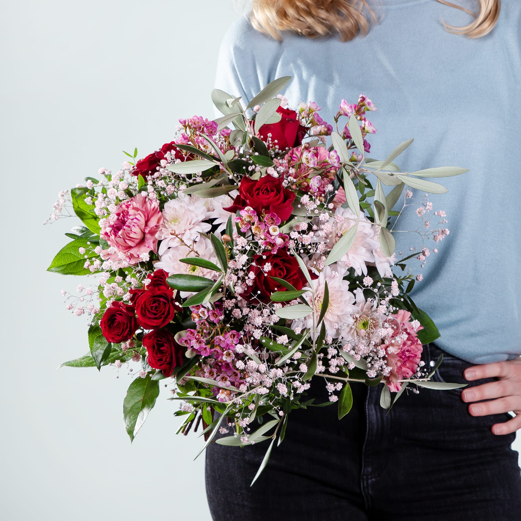 Ein leuchtender Strauß roter Rosen, rosa Nelken und anderer bunter Blumen wird vor einer Person gehalten, die einen hellblauen Pullover vor einem schlichten weißen Hintergrund trägt.
