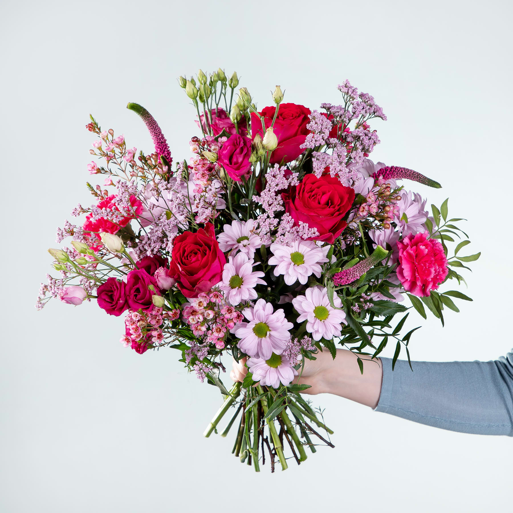 Ein leuchtender Strauß roter Rosen, rosa Nelken und weißer Gänseblümchen, den eine Person vor einem schlichten Hintergrund in den Händen hält.