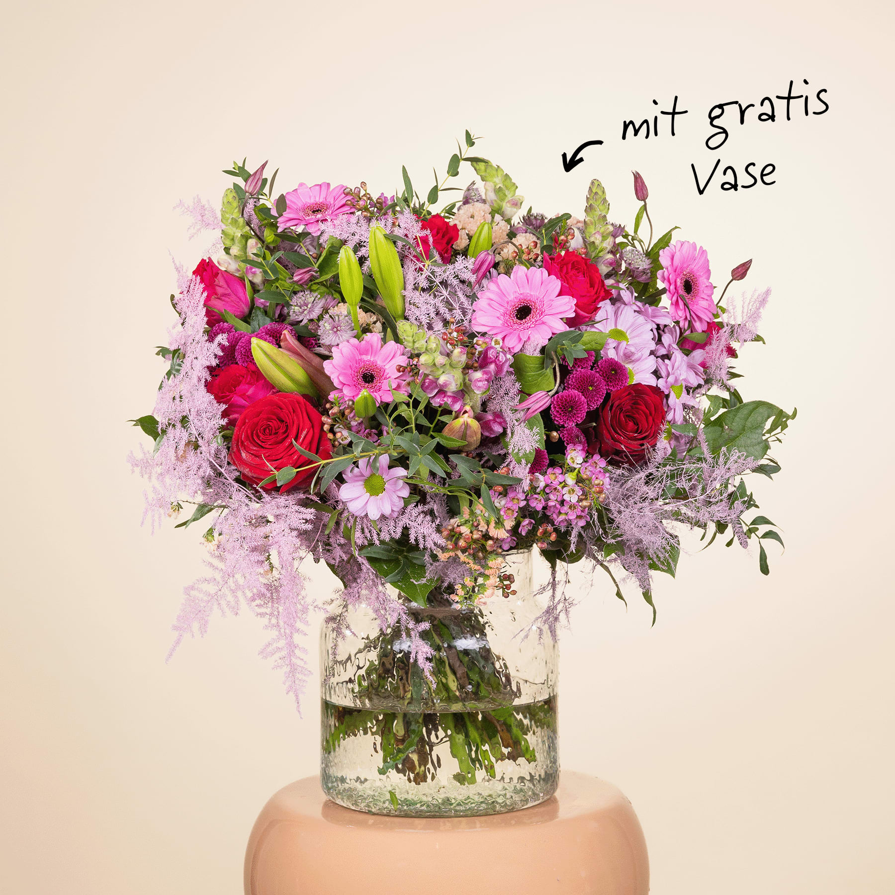 Ein lebendiger Strauß verschiedener bunter Blumen, darunter rosa Gerbera, rote Rosen sowie violette und weiße Blumen, wird in einer Glasvase vor einem schlichten beigen Hintergrund präsentiert.