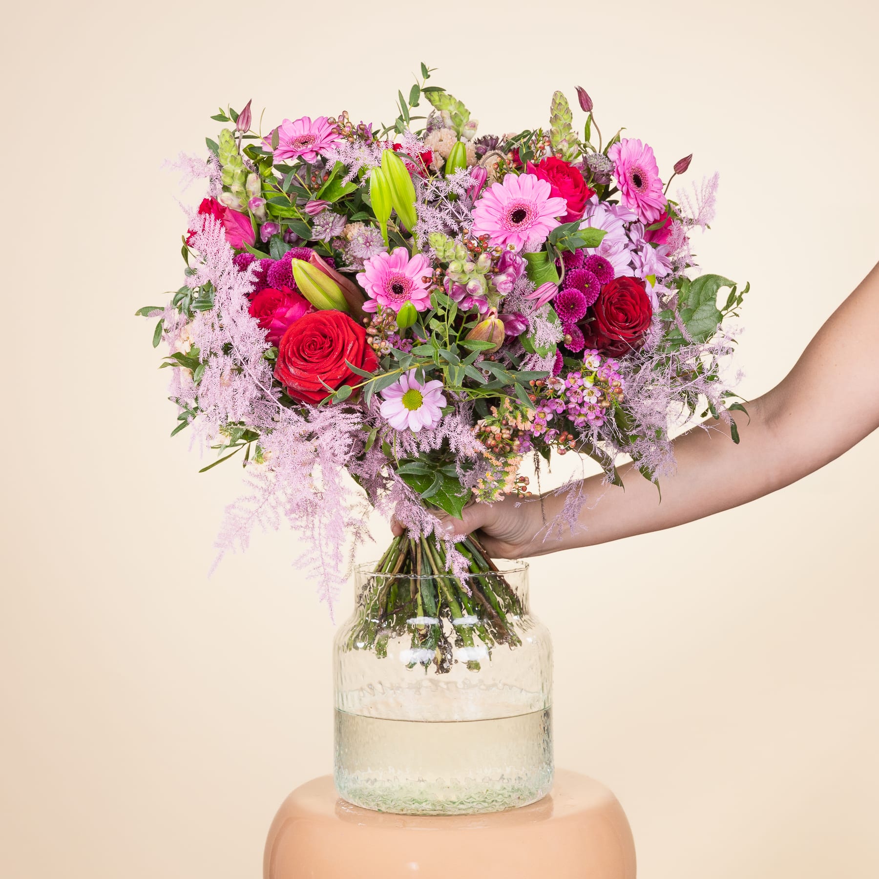 Ein farbenfroher Strauß rosa und roter Blumen, darunter Rosen und Gerbera, steht in einer Glasvase vor einem schlichten beigen Hintergrund.