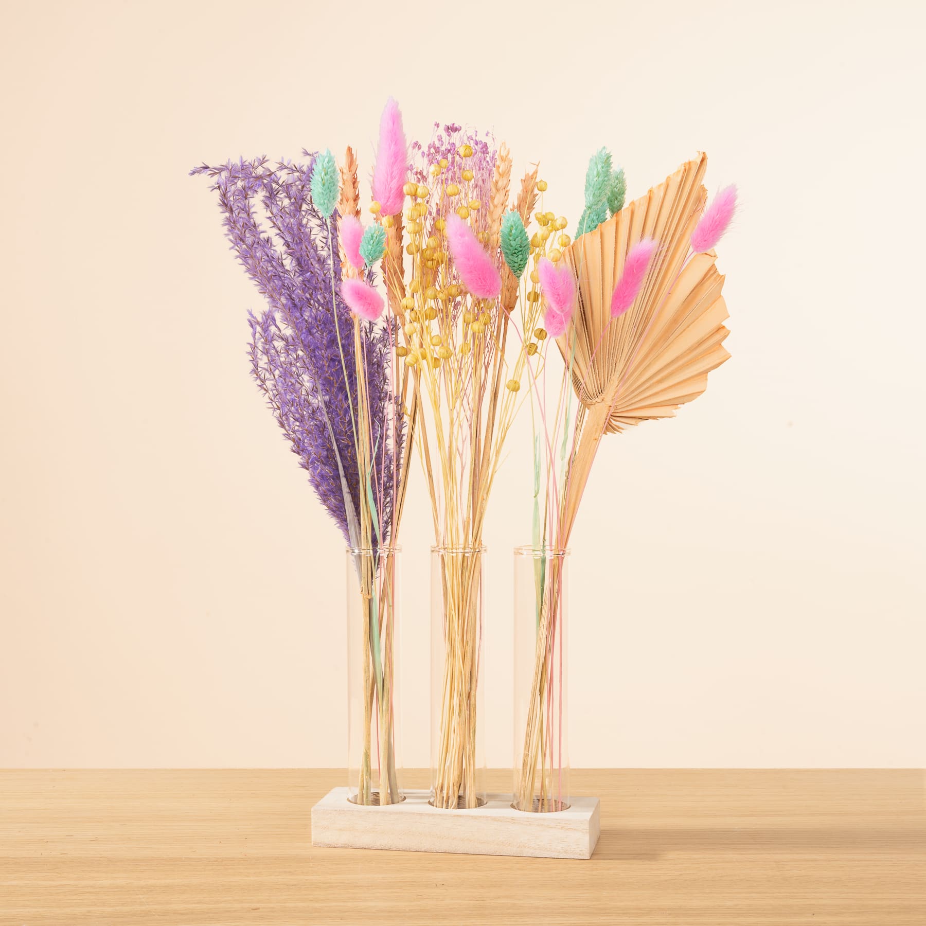 Ein Strauß bunter getrockneter Blumen und Pflanzen, darunter Lavendel, Pampasgras und Palmblätter, arrangiert in Glasvasen auf einer Holzoberfläche vor einem hellbeigen Hintergrund.