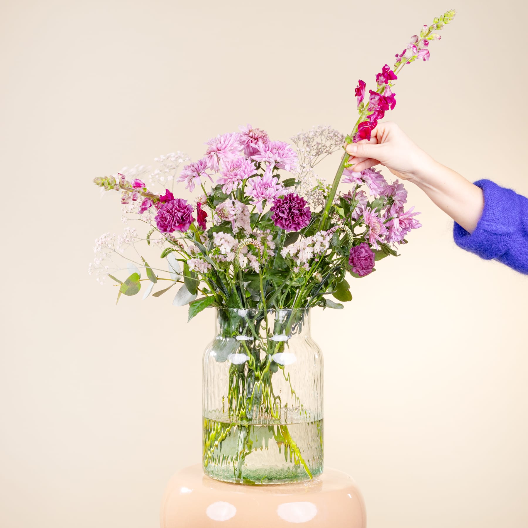 Ein Strauß leuchtend rosa und violetter Blumen, darunter Nelken und Löwenmäulchen, steht in einem Glasgefäß vor einem schlichten, hellen Hintergrund.