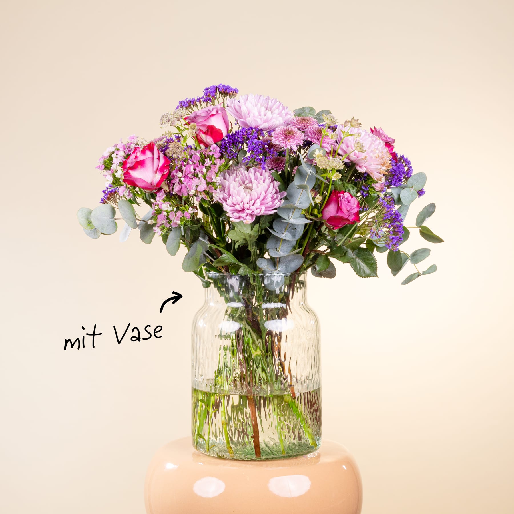 Ein lebendiger Strauß verschiedener bunter Blumen, darunter rosa Rosen, violette Astern und grünes Laub, wird in einer Glasvase vor einem schlichten, hellen Hintergrund präsentiert.