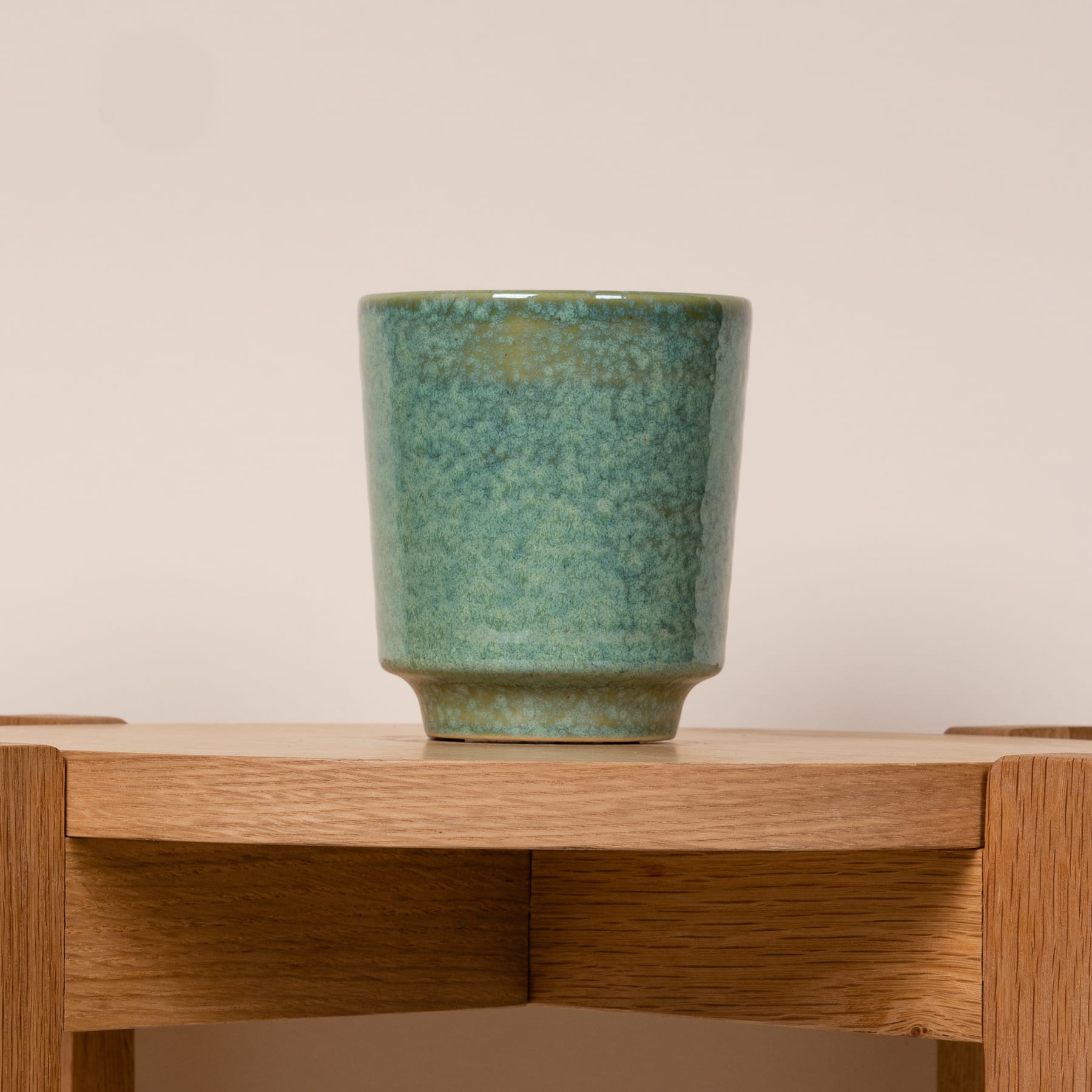 Eine grüne Keramiktasse oder -vase steht auf einem Holzregal oder -tisch vor einem hellen Hintergrund.
