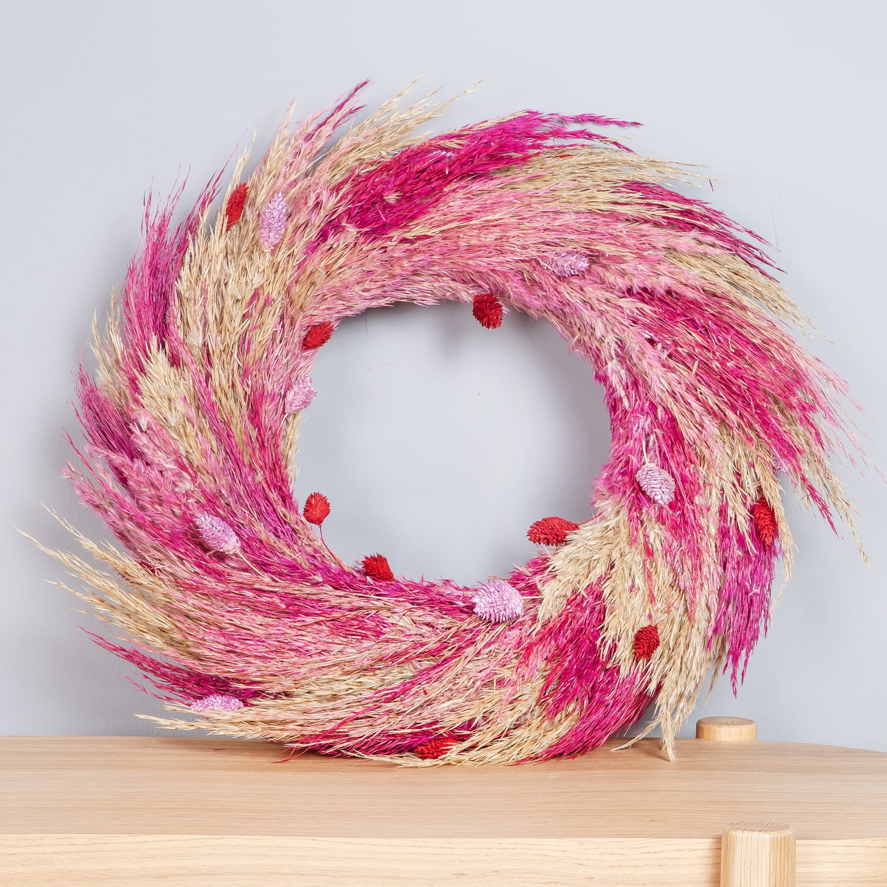 Ein leuchtender, runder Kranz aus getrockneten Gräsern oder Schilf in Rosa und Beige liegt auf einer Holzoberfläche vor einem schlichten weißen Hintergrund.