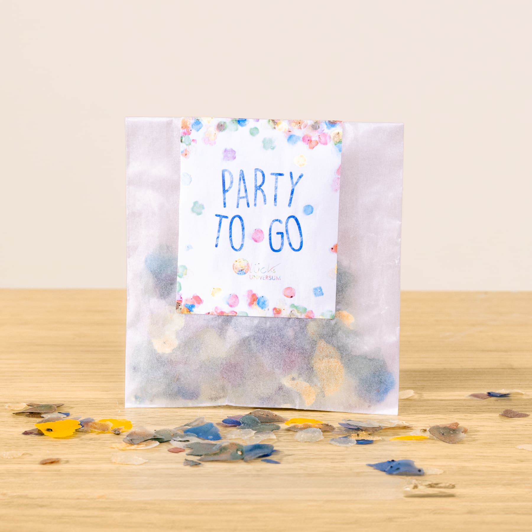 Auf einer Holzfläche steht eine durchsichtige Plastiktüte gefüllt mit buntem Konfetti, davor klebt eine Karte mit der Aufschrift „Party to go“.
