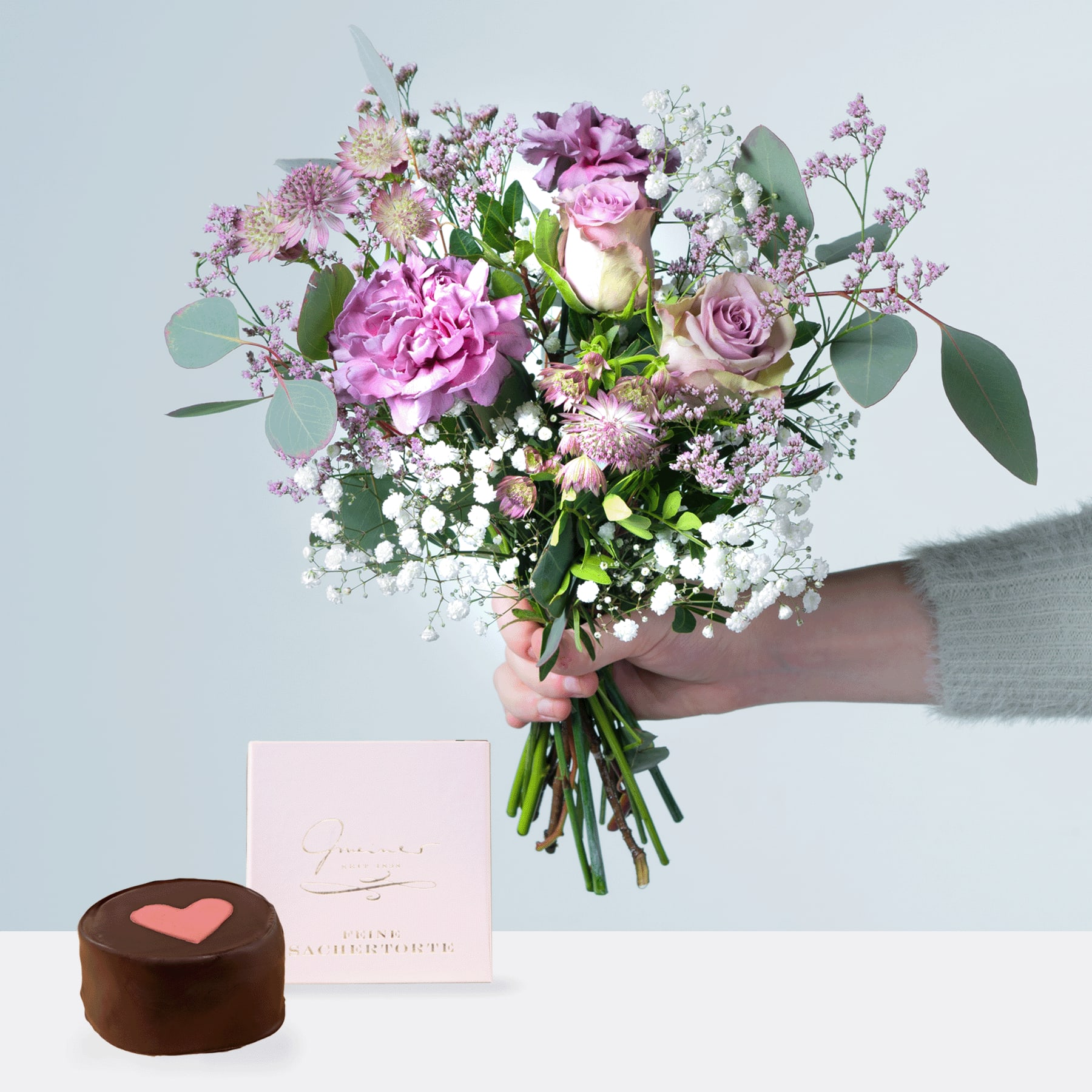 Eine Person hält einen farbenfrohen Strauß rosa und violetter Blumen, darunter Rosen und Pfingstrosen, in den Händen, begleitet von einer kleinen herzförmigen Schokolade auf einer rosa Karte.