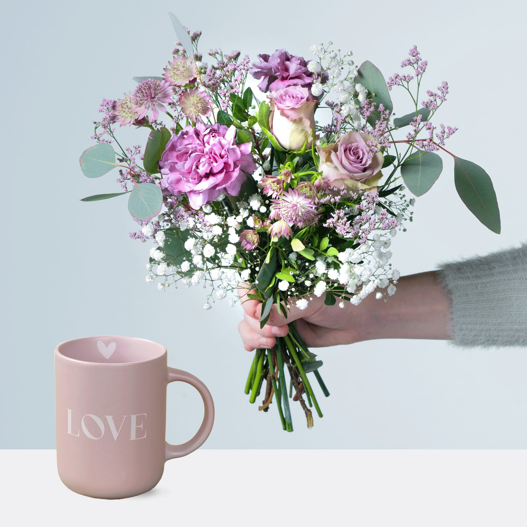 Ein Strauß leuchtend rosa und violetter Blumen, darunter Rosen und Pfingstrosen, wird vor einem pastellrosa Becher gehalten, auf dem vor einem hellblauen Hintergrund das Wort „LOVE“ steht.