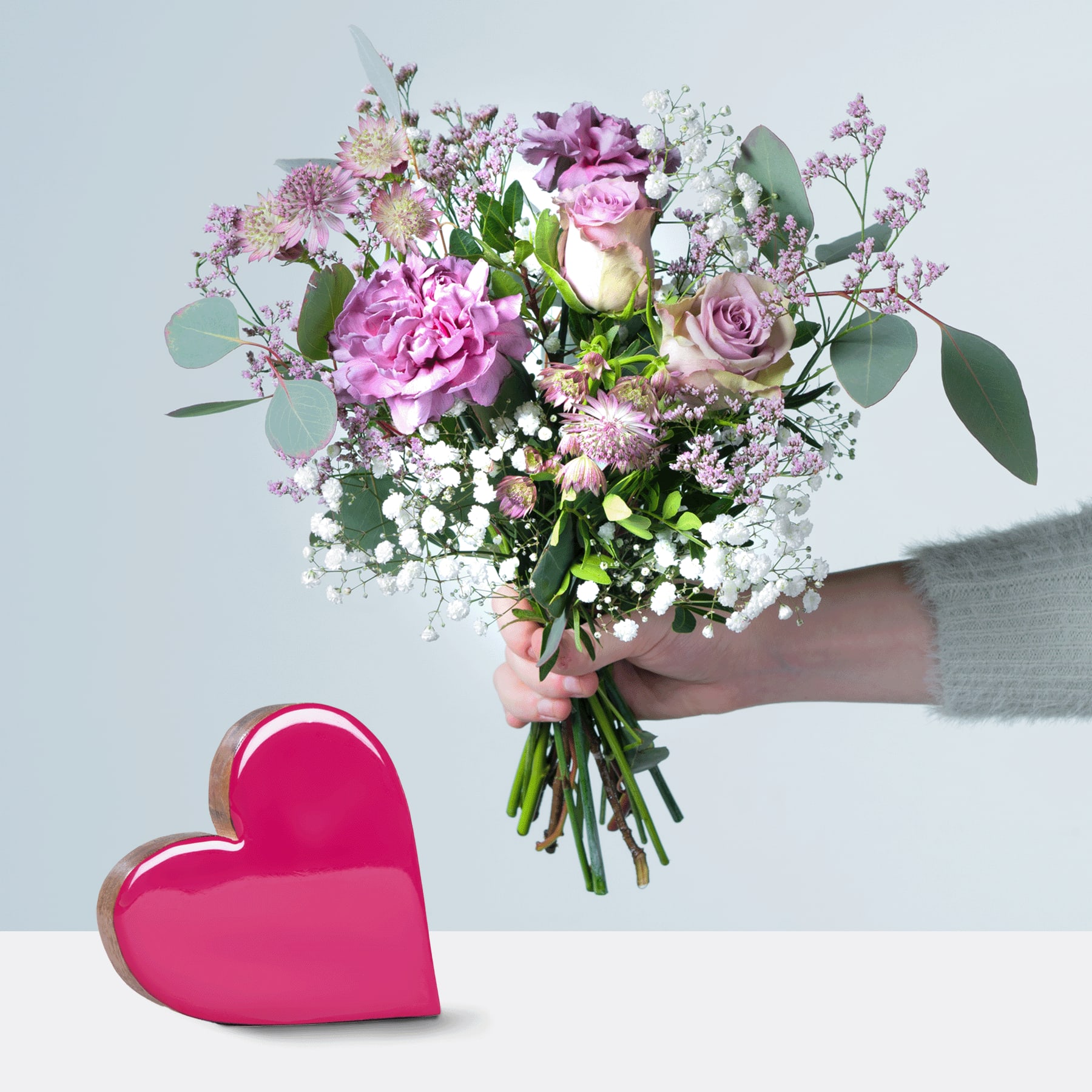 Eine Person hält einen farbenfrohen Strauß rosa und violetter Blumen, darunter Rosen und Pfingstrosen, in den Händen. Im Vordergrund steht auf einer weißen Oberfläche eine rote herzförmige Schachtel.