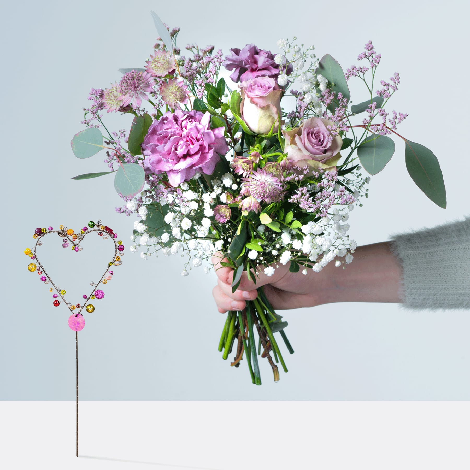 Ein farbenfroher Strauß rosa und violetter Blumen, darunter Rosen und Pfingstrosen, wird vor einem schlichten grauen Hintergrund in den Händen einer Person gehalten, im Vordergrund ist eine herzförmige Drahtdekoration zu sehen.