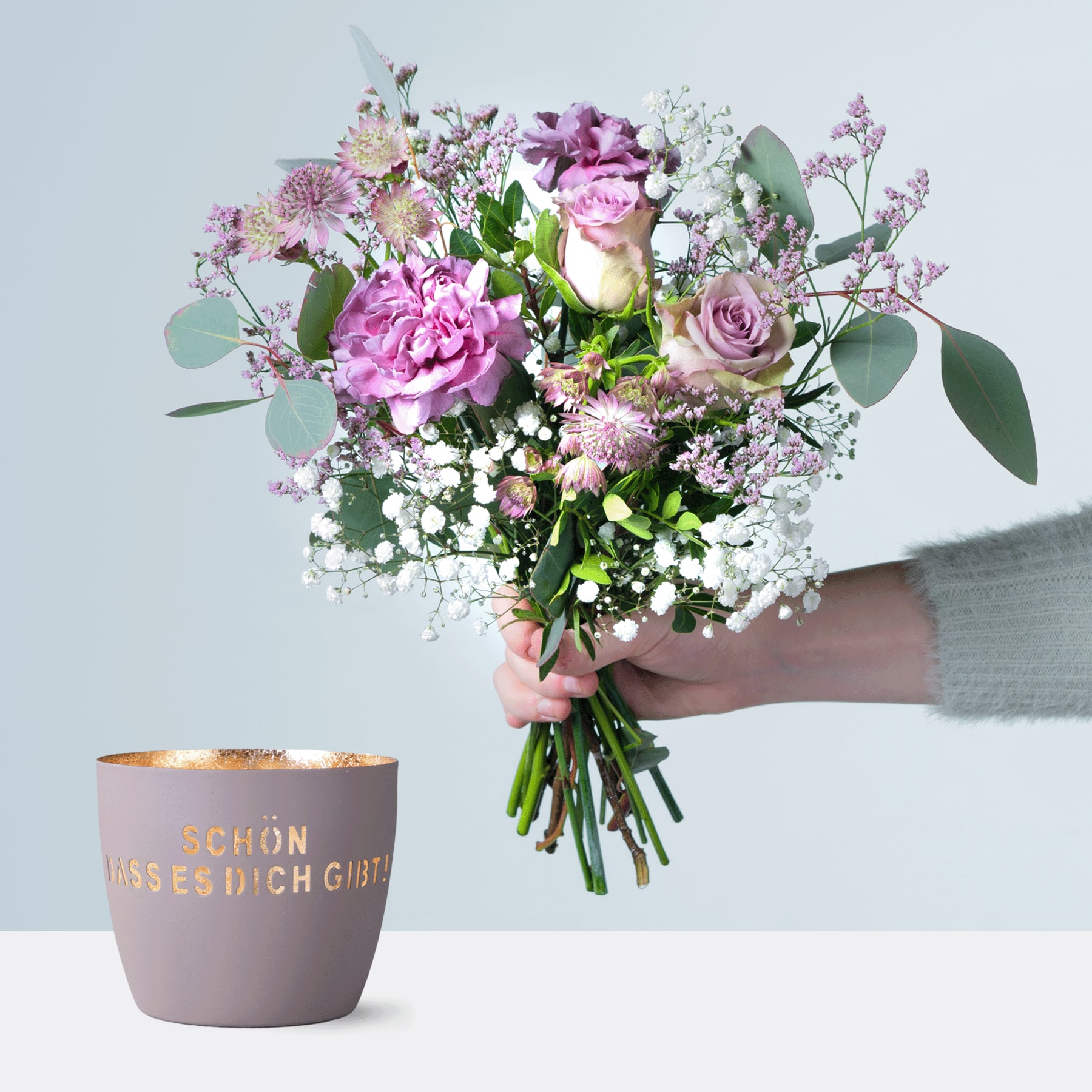 Eine Person hält einen farbenfrohen Strauß rosa und violetter Blumen, darunter Rosen und Pfingstrosen, in den Händen, begleitet von einer Keramiktasse mit deutschem Text auf hellem Hintergrund.