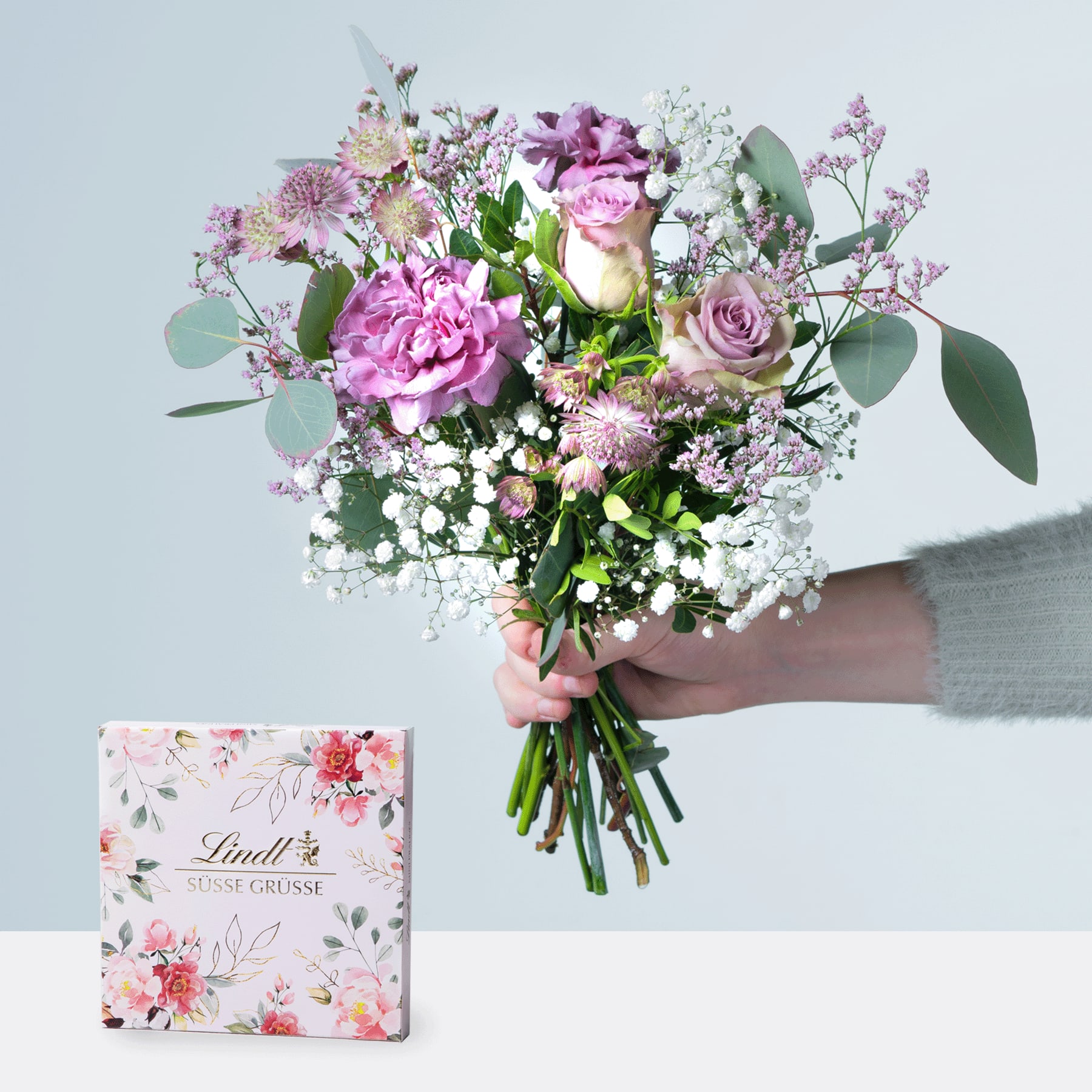 Eine Person hält einen farbenfrohen Strauß rosa und violetter Blumen, darunter Rosen und Pfingstrosen, in den Händen, begleitet von einer Schachtel Lindt-Pralinen auf einer weißen Oberfläche.