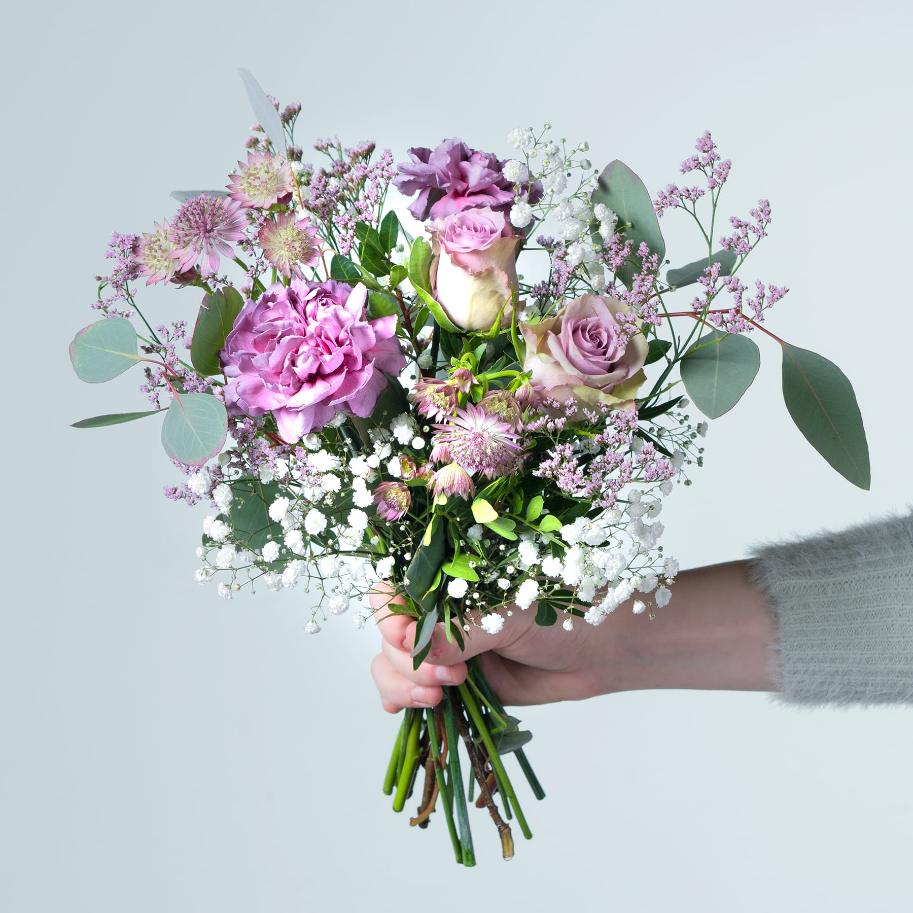 Ein leuchtender Strauß rosa und violetter Blumen, darunter Pfingstrosen, Rosen und andere zarte Blüten, wird vor einem schlichten, hellen Hintergrund in den Händen einer Person gehalten.